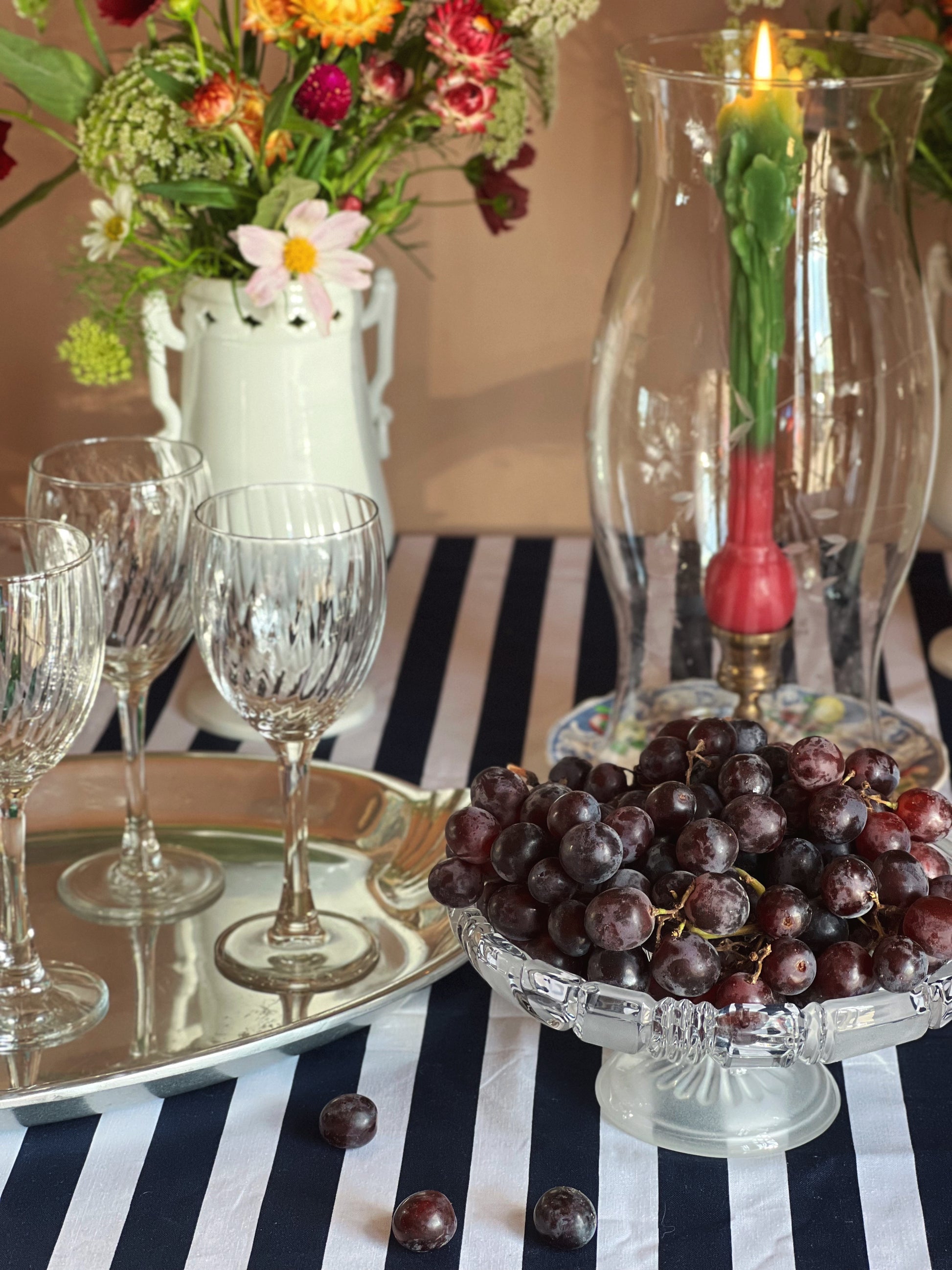 A styled tabletop scene with a glass pedestal bowl filled with dark purple grapes, a silver tray with clear stemmed glasses, a white pitcher of mixed flowers, and a tall clear glass candle holder with a lit green taper and red top, all set on a navy-and-white striped tablecloth.