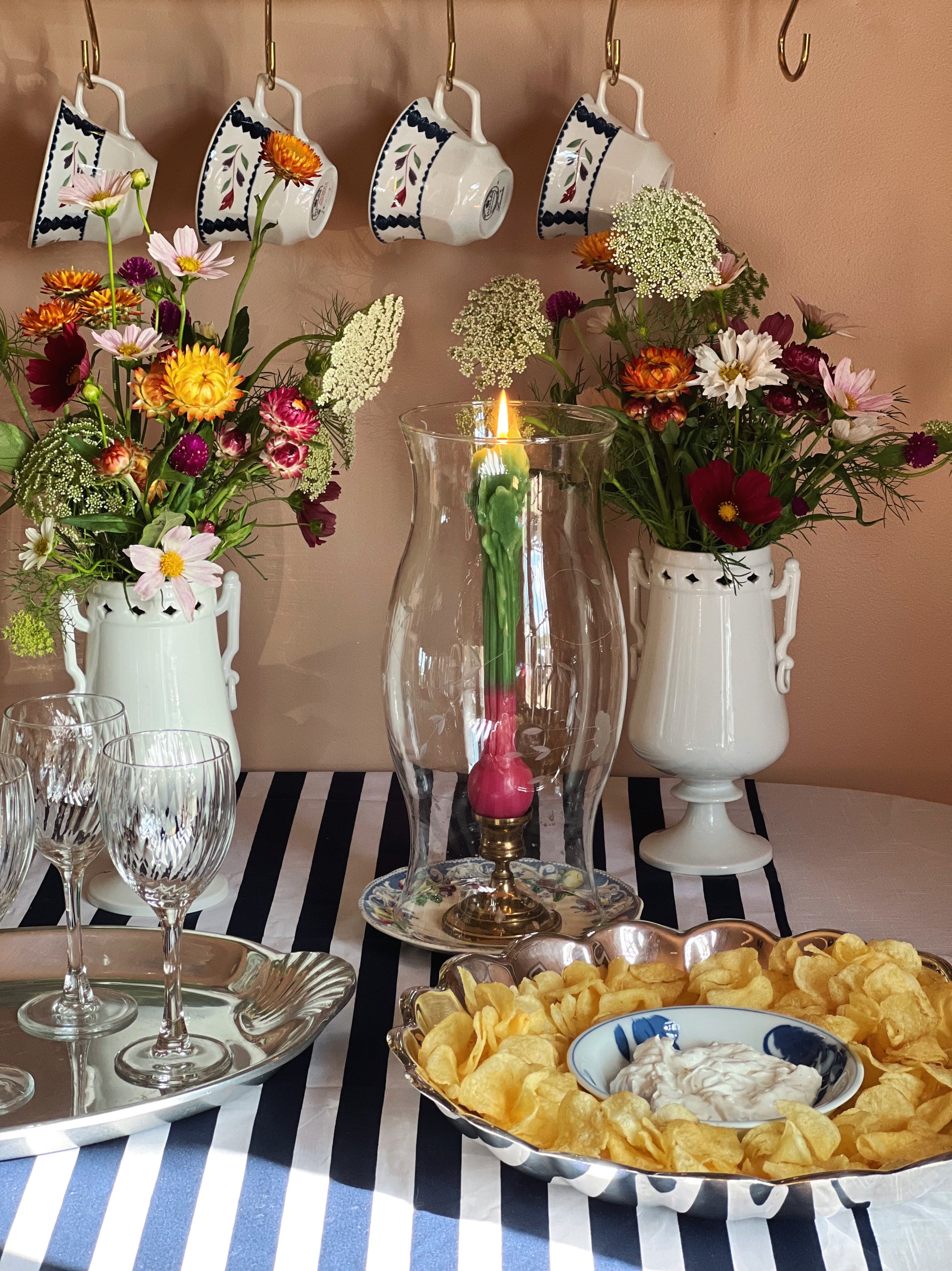 A styled tabletop scene with a blue-and-white striped tablecloth, a vintage Reed & Barton Holiday silverplate chip and dip serving bowl with potato chips and onion dip, two white pitchers filled with mixed flowers, and a tall clear glass vase with a lit candle in the center. Decorative teacups hang on the wall above.