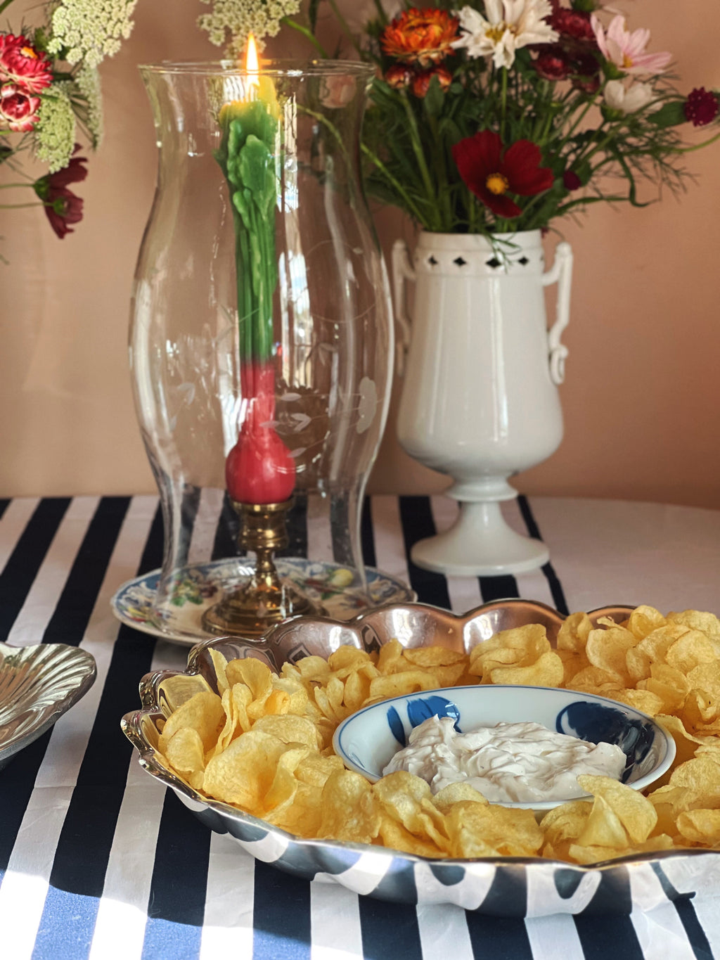 A styled tabletop scene with a Vintage Reed & Barton Holiday silverplate chip and dip serving bowl with scalloped edge styled with potato chips and onion dip, set on a navy-and-white striped cloth. In the background are two tall glass candle holders with lit candles and floral arrangements in ceramic vases.