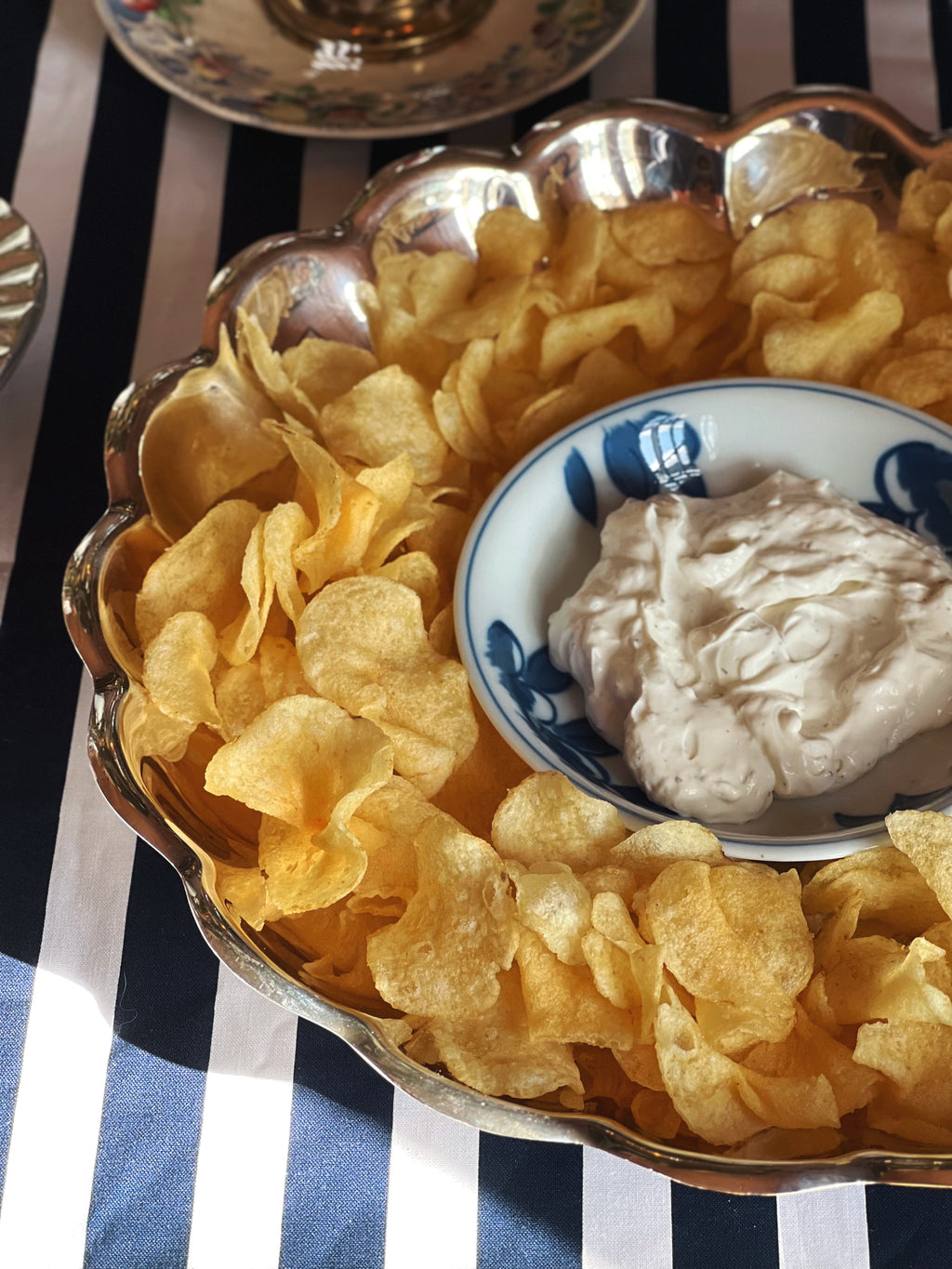 A scalloped silver serving platter filled with golden potato chips and a small blue-and-white bowl of creamy dip, set on a navy-and-white striped tablecloth.