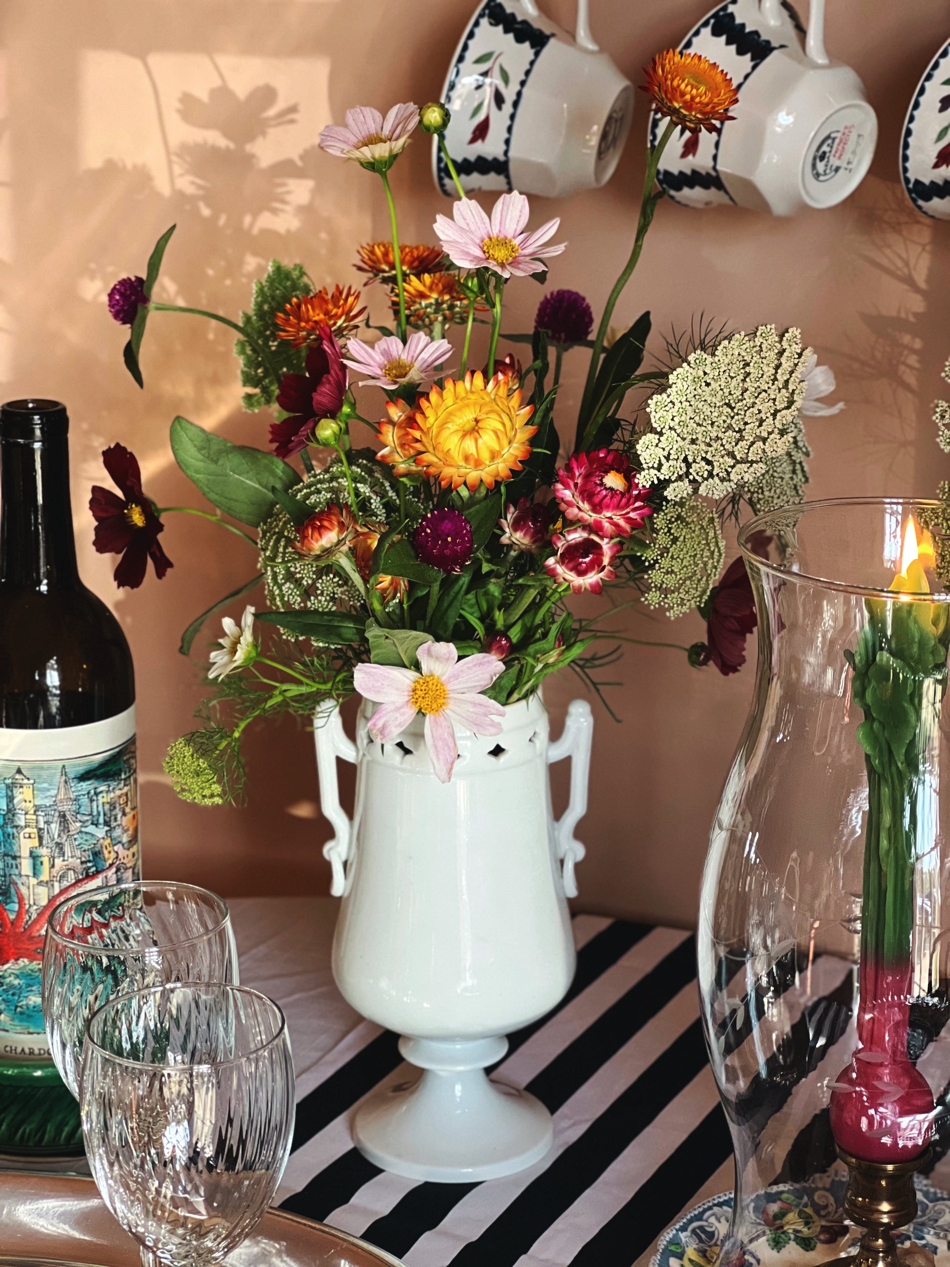 A white ceramic pedestal vase filled with a colorful wildflower arrangement, styled on a table with a striped runner, crystal glasses, a wine bottle, and hanging teacups against a warm beige wall.
