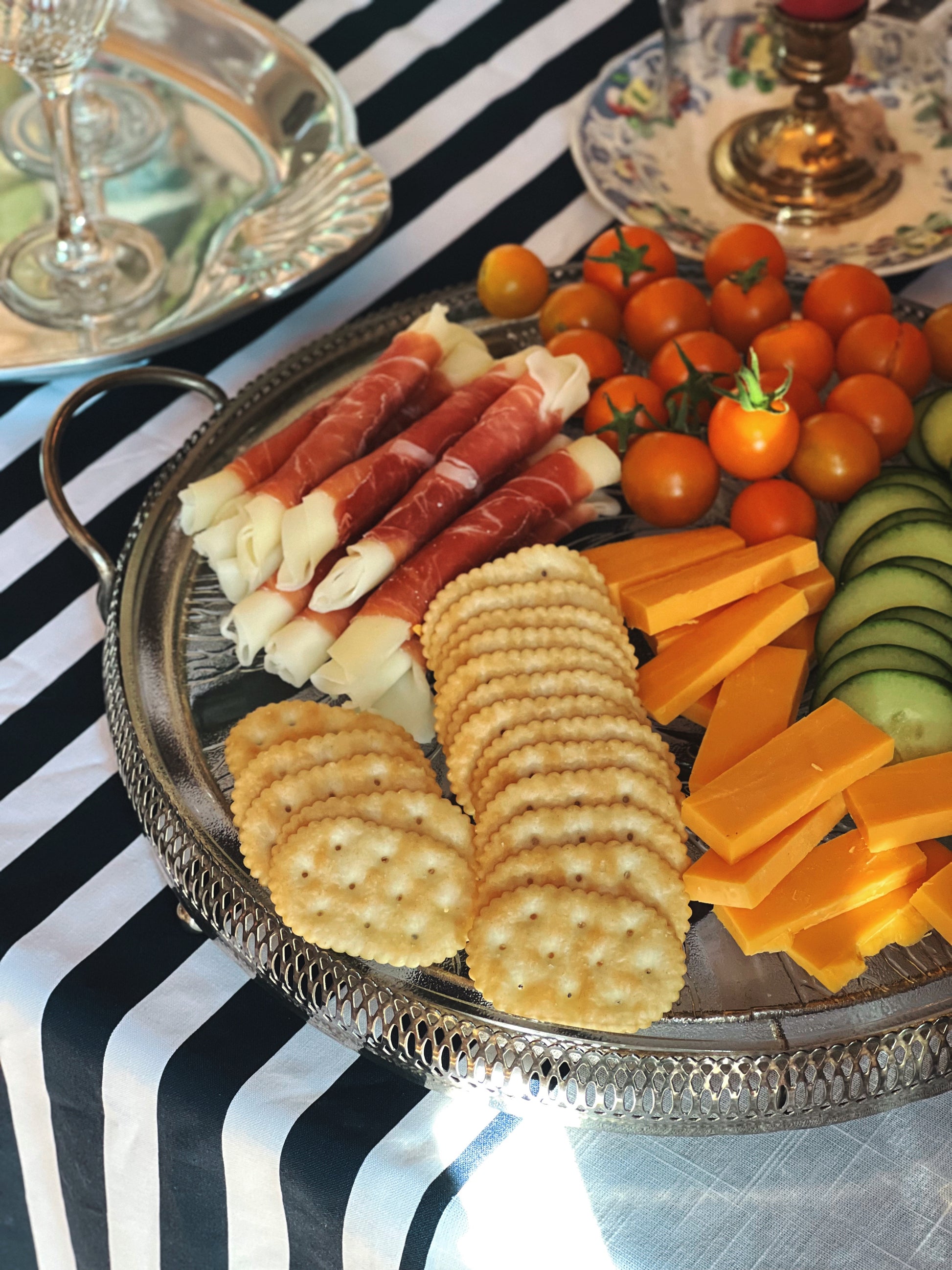 Vintage frosted swirl glass serving platter in a silver footed stand styled with charcuterie, crackers, cheese, grapes, and tomatoes styled on a table with a striped tablecloth.