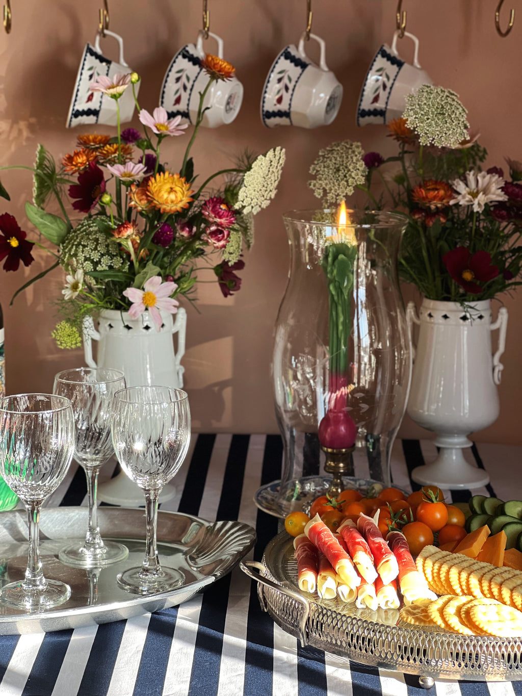 A styled tabletop scene with floral arrangements in white urns, hanging teacups against a peach wall, wine glasses on a silver tray, and charcuterie-style food on a wire platter over a blue-and-white striped tablecloth.