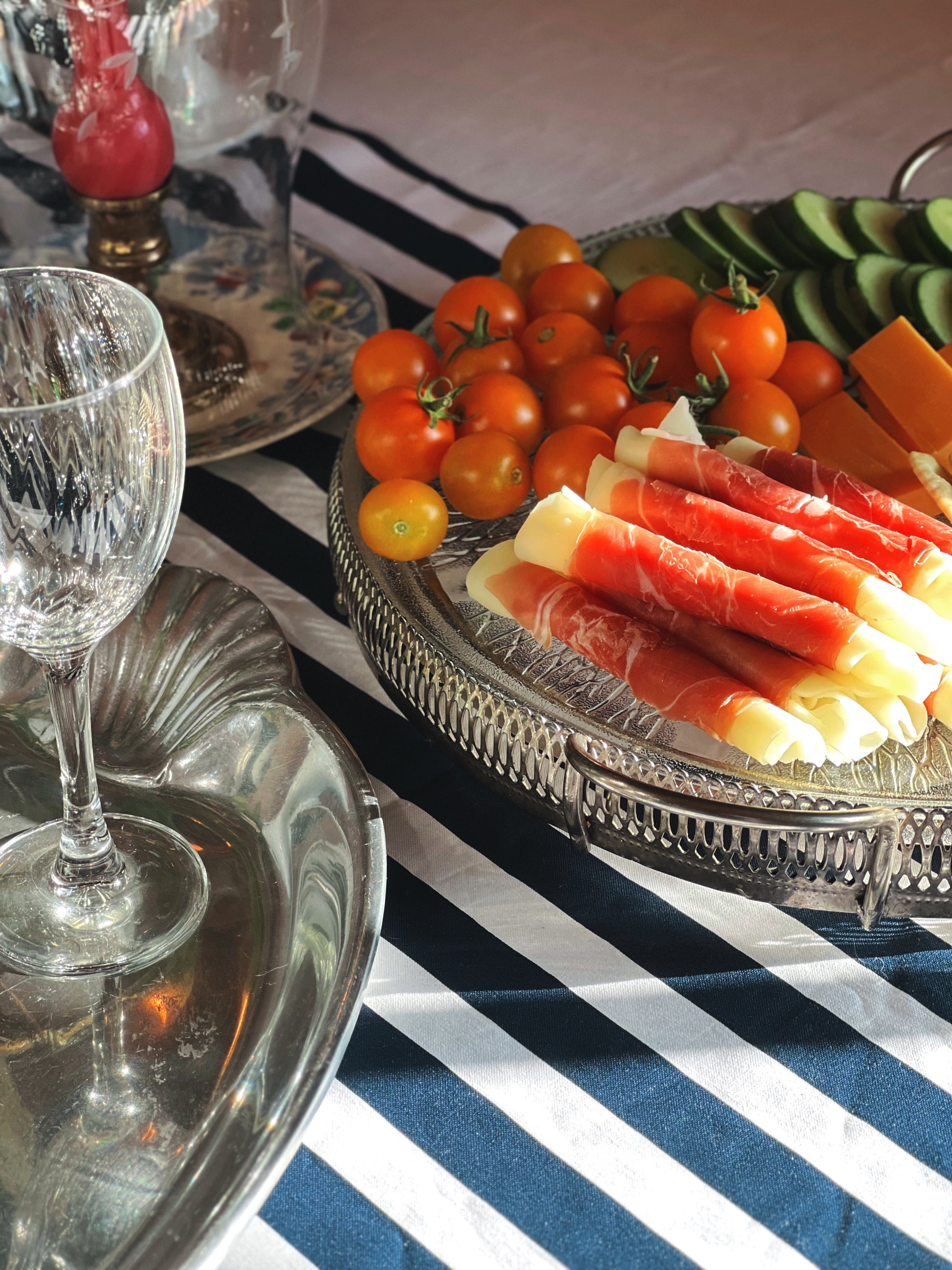 A close-up of a silver serving platter filled with sliced cured meats, cherry tomatoes, and cheese, set on a blue and white striped tablecloth next to a wine glass and silver tray.