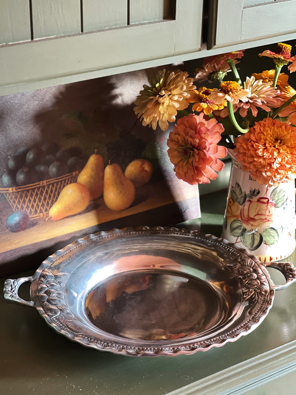 A round silver serving platter with ornate handles and a scalloped rim, displayed on a tabletop next to a floral arrangement and a framed still life painting.