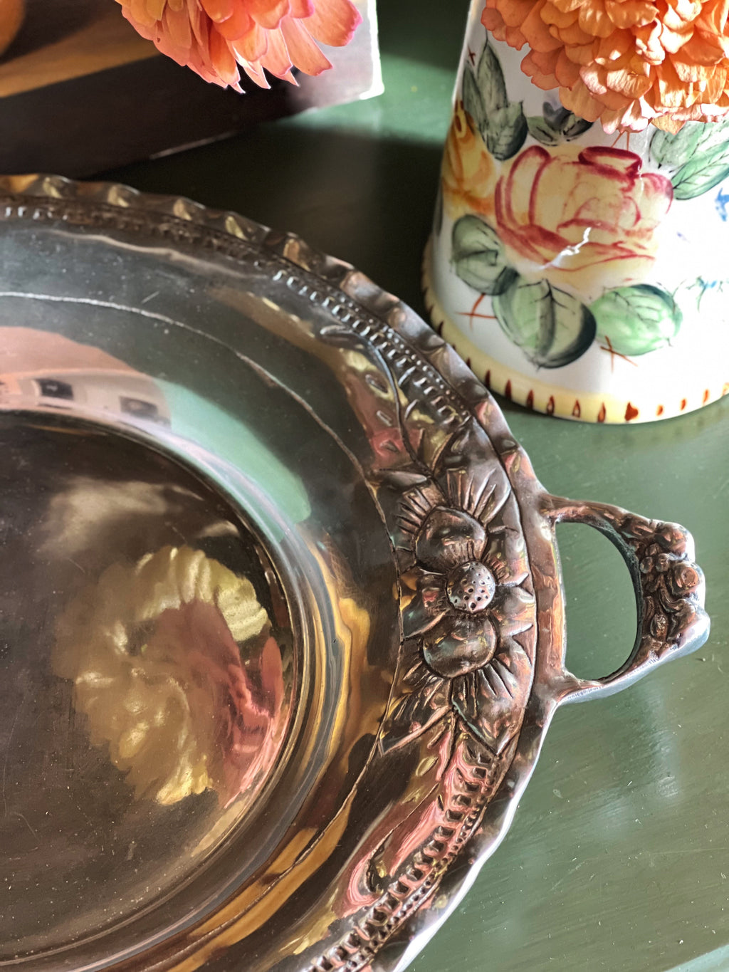 A close-up of a decorative silver-toned metal tray with embossed floral detailing and a side handle, placed on a dark green surface next to a floral ceramic vase.