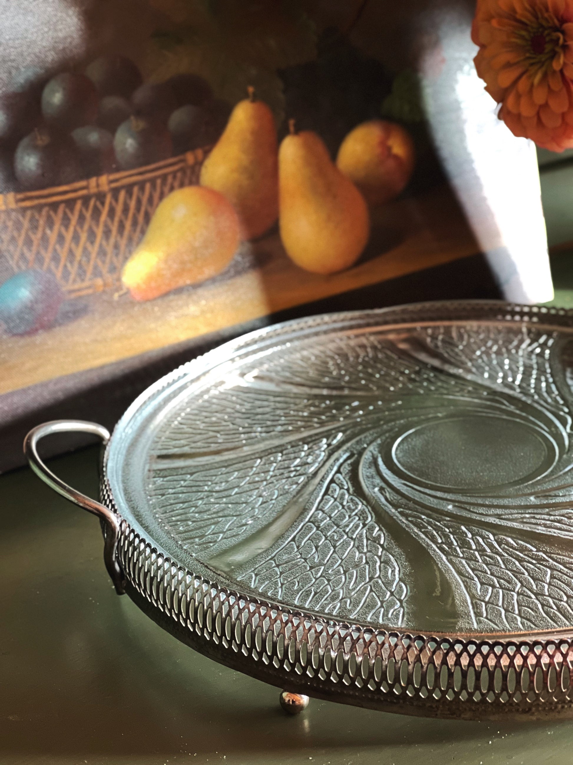 A round silver metal serving tray with a decorative embossed swirl pattern and a fluted rim, shown on a tabletop with a blurred fruit still life in the background.