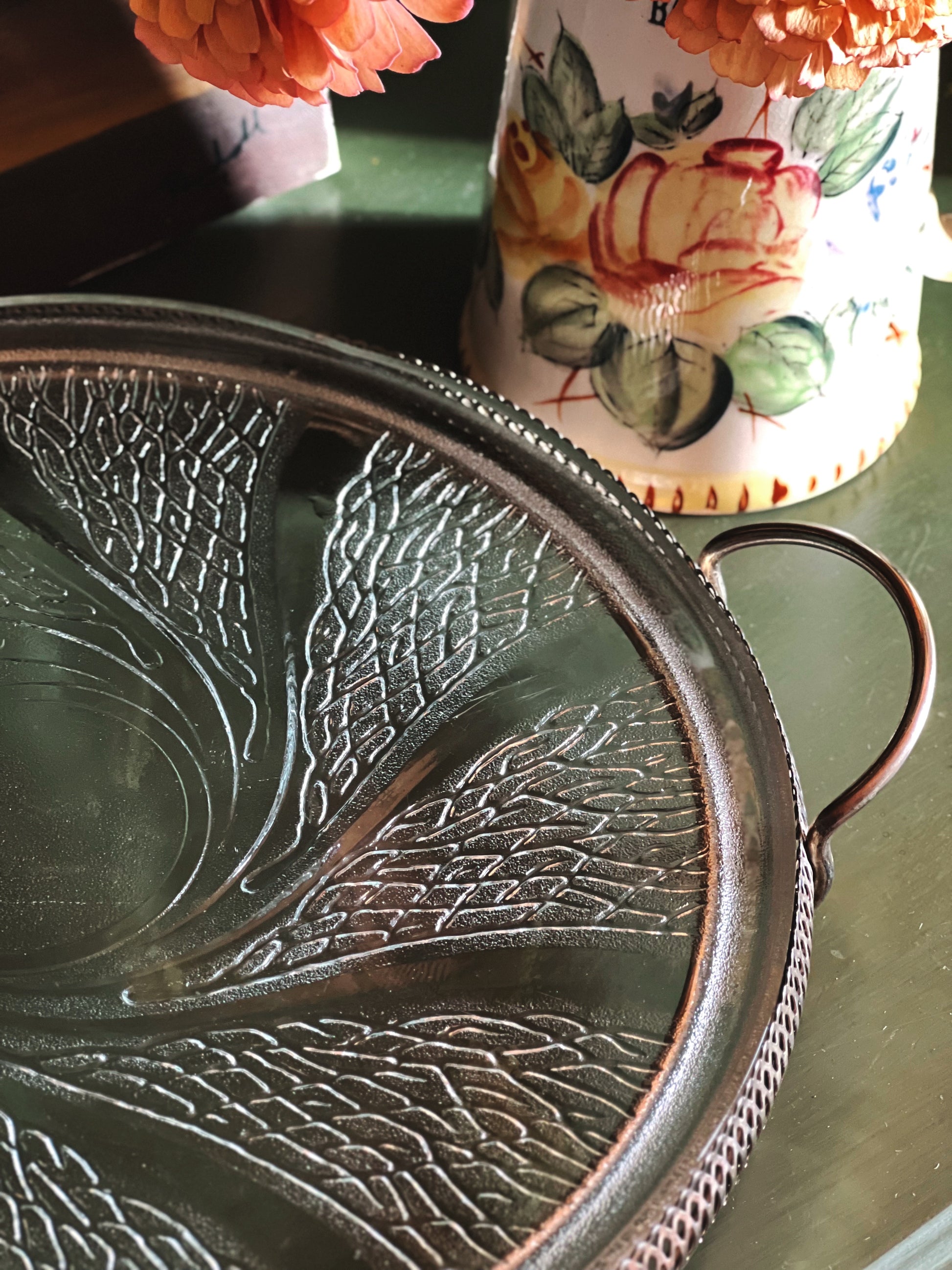 A close-up of a Vintage frosted swirl glass serving platter in a silver footed stand styled set on a dark tabletop with a floral ceramic vase in the background.
