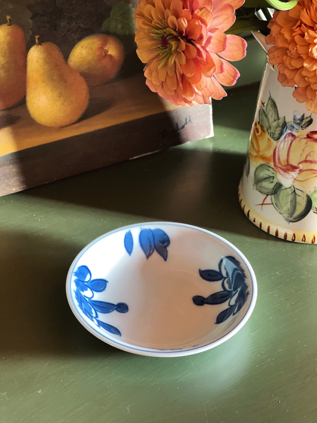 A small white ceramic bowl with blue floral patterns, placed on a green tabletop next to a floral vase and a painted still life of pears.