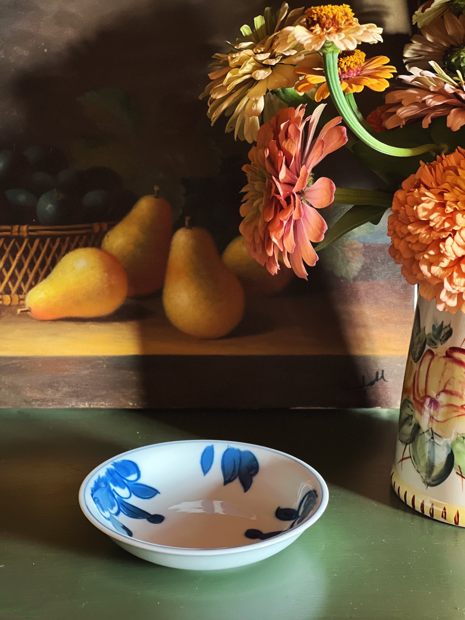 A small white ceramic bowl with blue floral patterns sits on a green table in the foreground, with a painted still life scene of pears and flowers behind it.