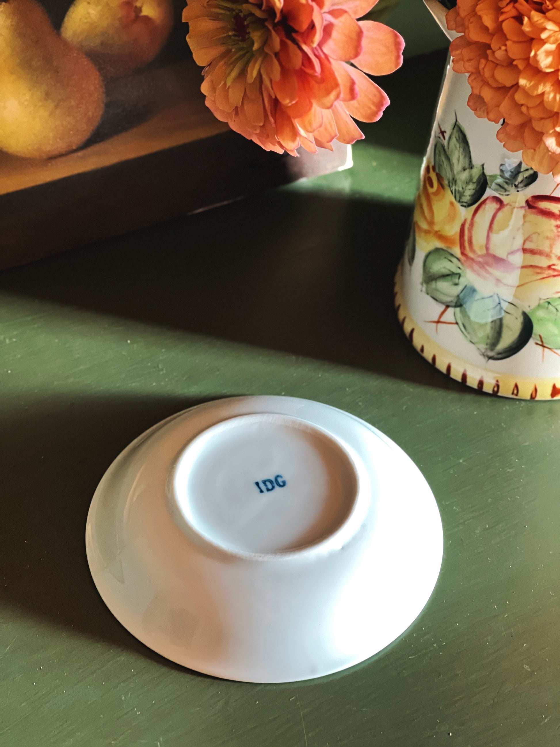 A white ceramic bowl or saucer shown upside down on a dark green tabletop, with the blue monogram 'IDG' printed inside the bottom. A floral-patterned vase with orange flowers and a fruit tray are partially visible in the background.