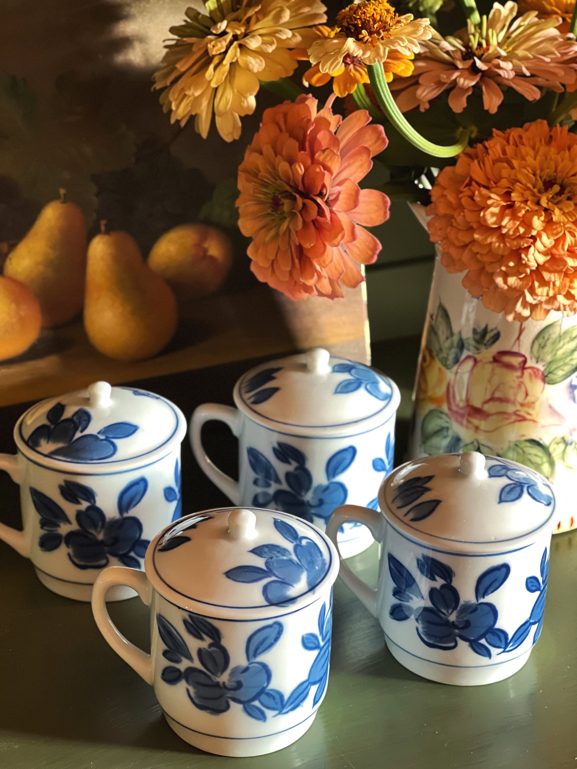 Four white ceramic lidded mugs with blue floral patterns arranged on a table, with a vase of orange flowers and pears in the background.