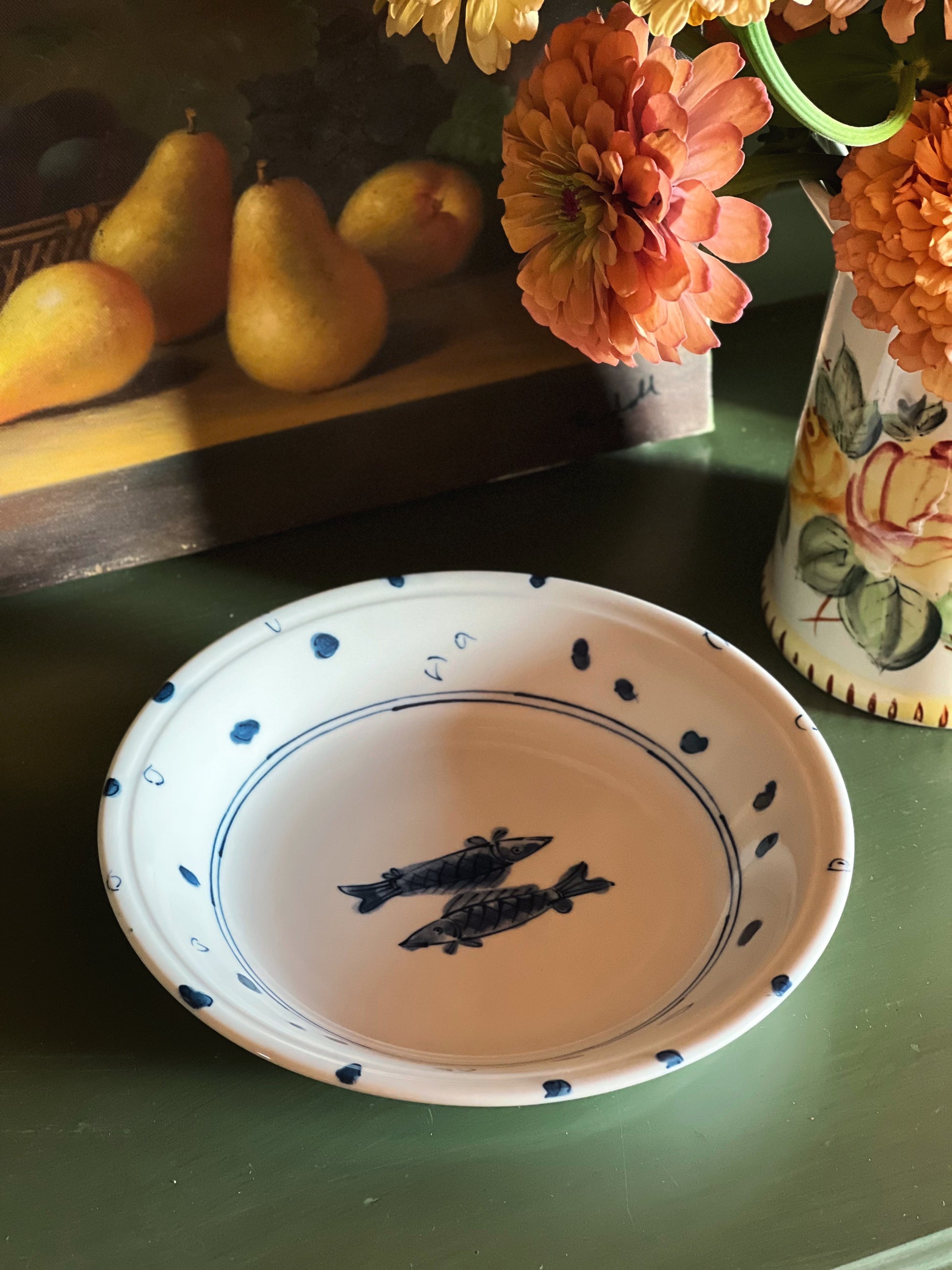Vintage blue and white porcelain serving bowl with fish design and dotted border on a green table beside flowers.