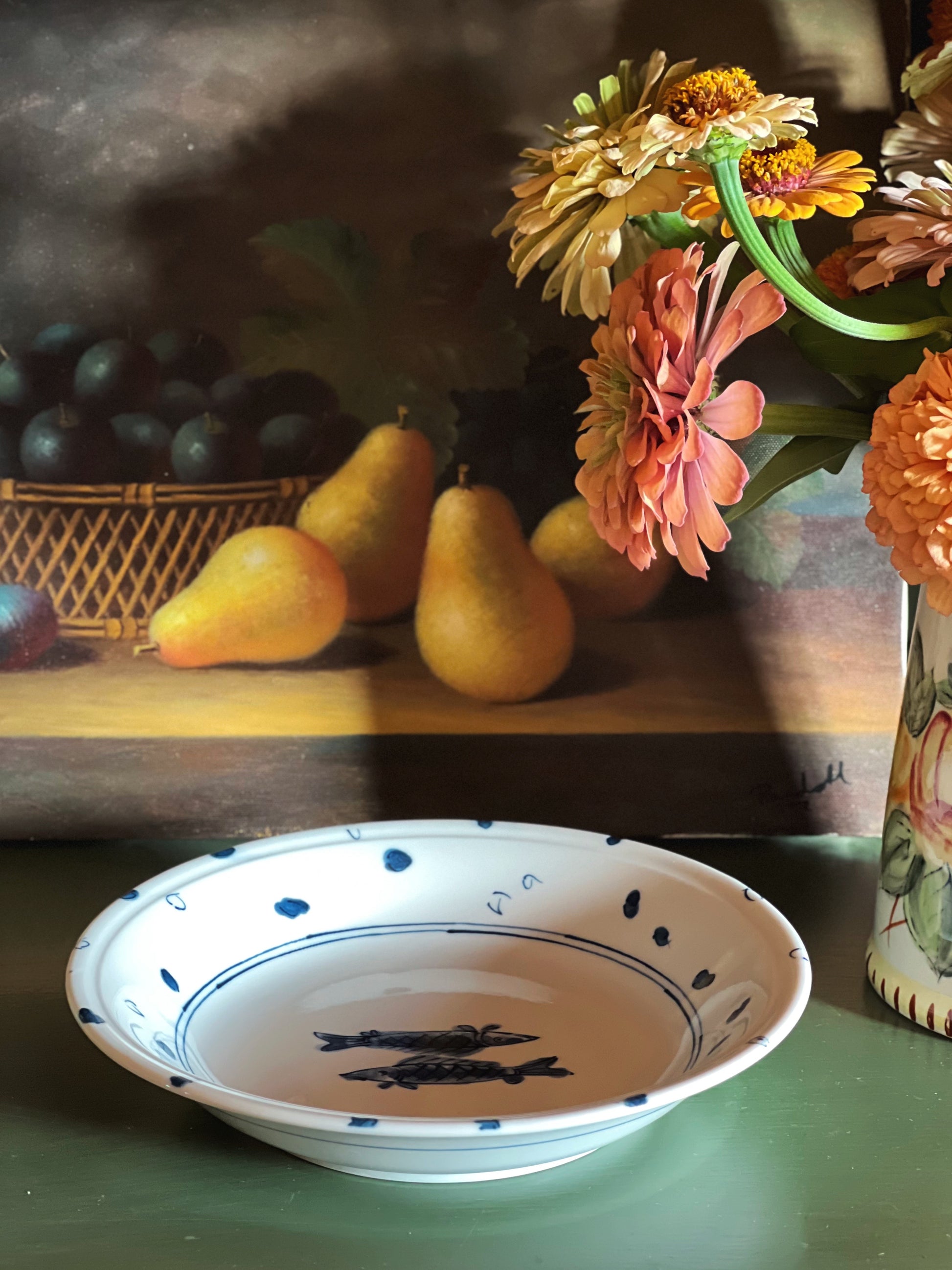 A white ceramic shallow bowl with blue painted dots and fish motifs, placed on a green tabletop in front of a painted still life of pears and flowers.