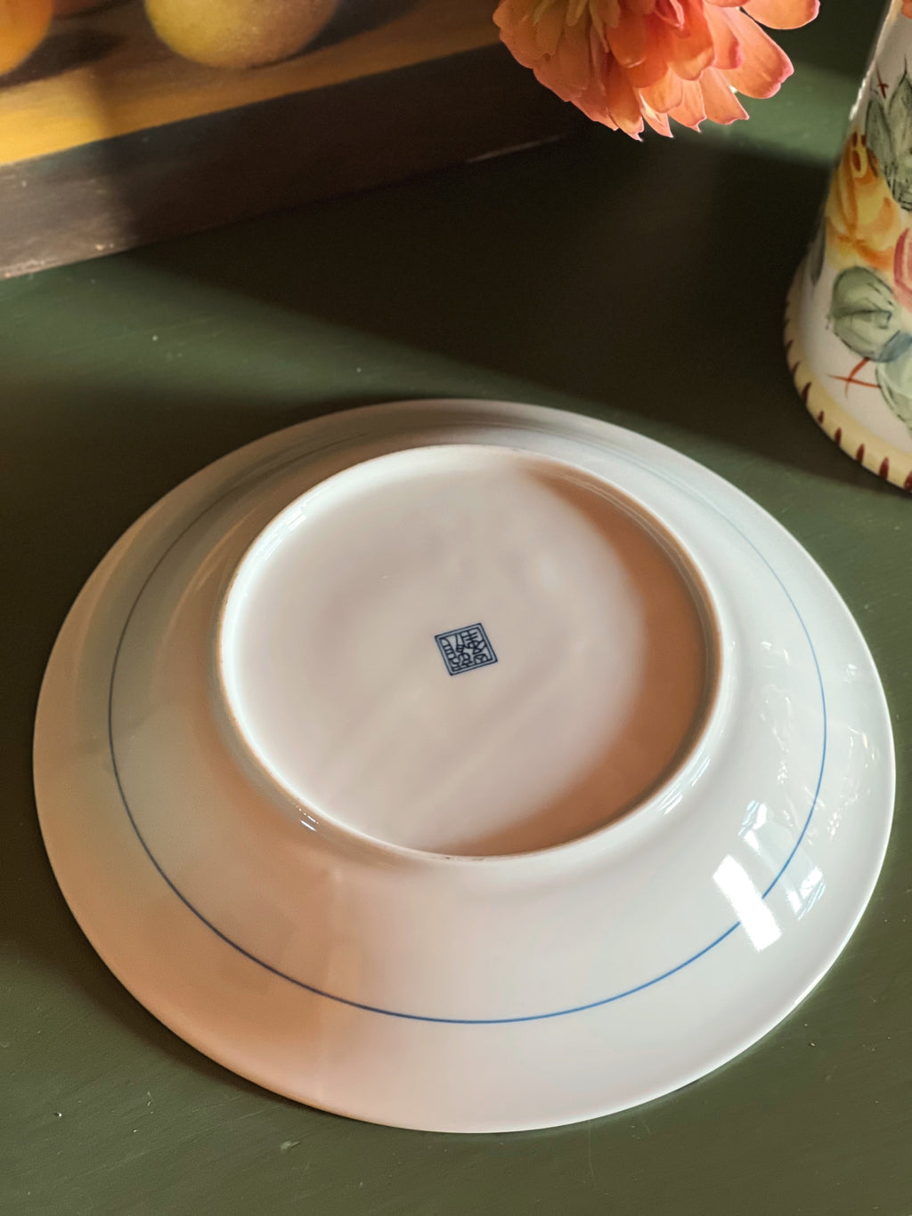 A white ceramic plate shown from the bottom, featuring a blue double-line border design and a small blue square mark in the center, resting on a dark green tabletop.