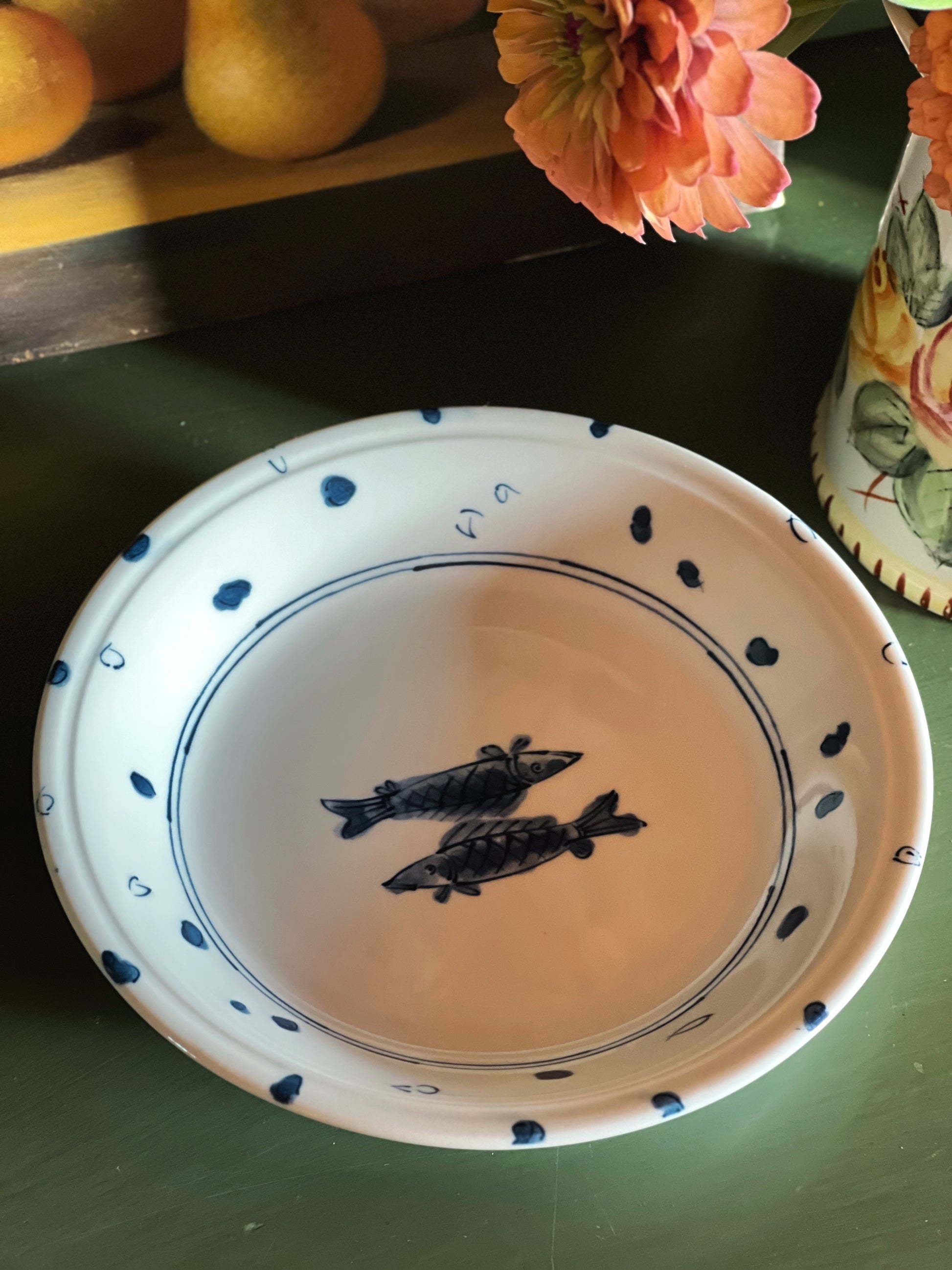 A white ceramic bowl with blue hand-painted fish and dots, shown on a dark green tabletop with flowers and fruit in the background.