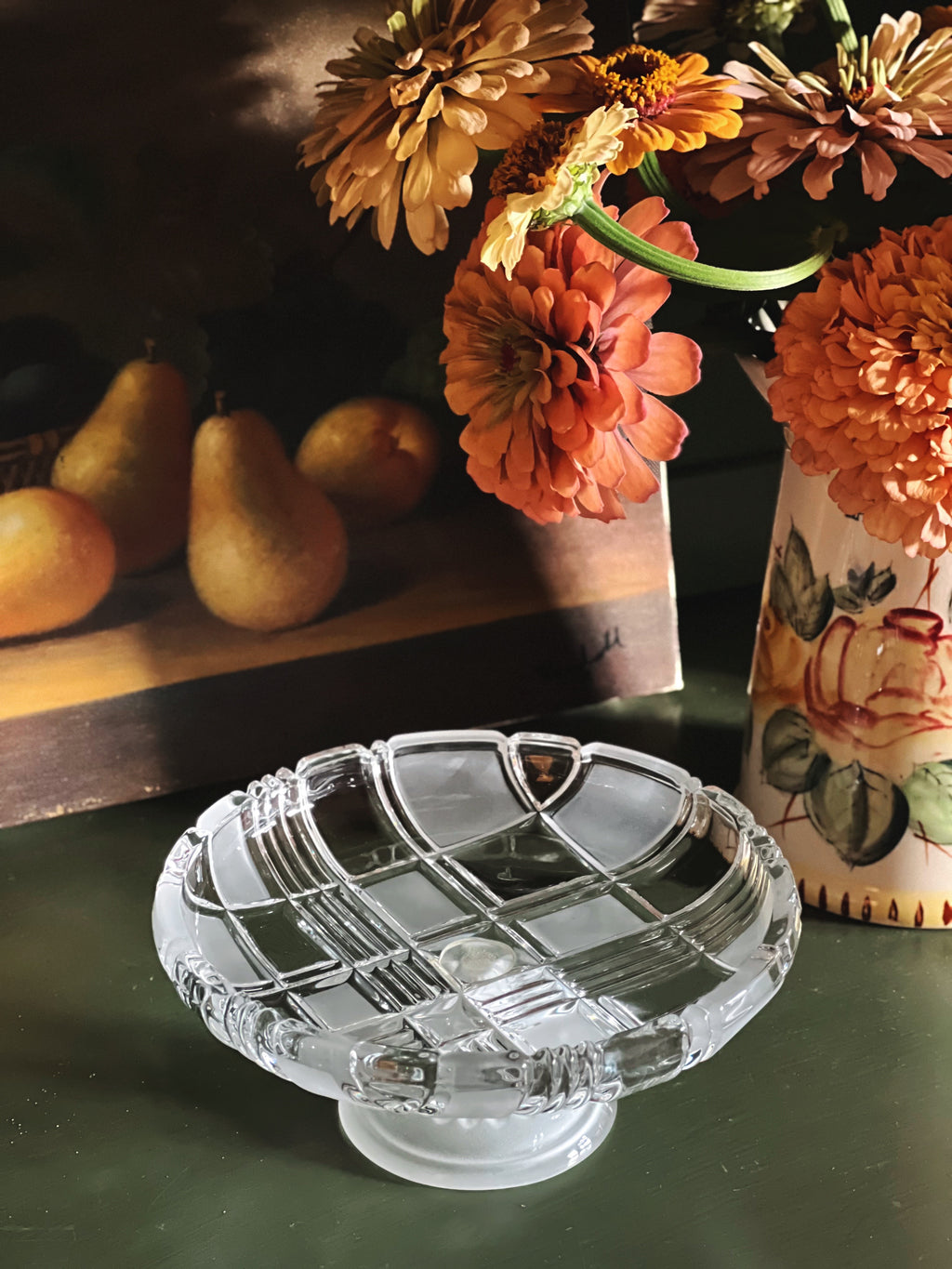 A clear glass pedestal plate with a grid pattern, set on a dark green table next to a floral vase and pears in the background.