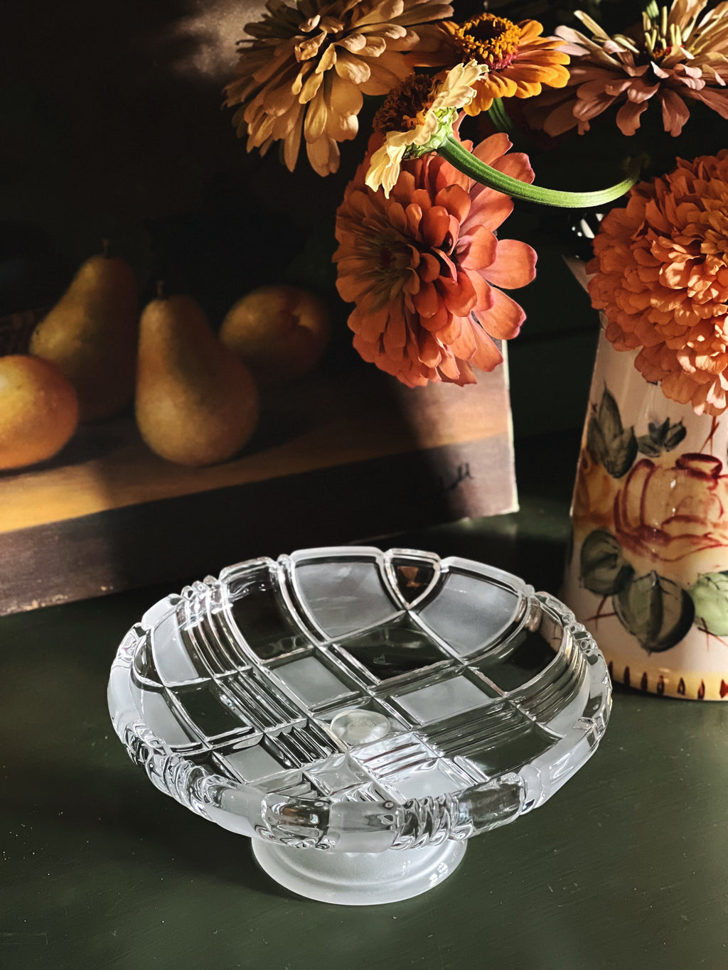A clear glass pedestal plate with a grid pattern and ribbed rim, set on a dark green surface next to a floral vase and pears in the background.