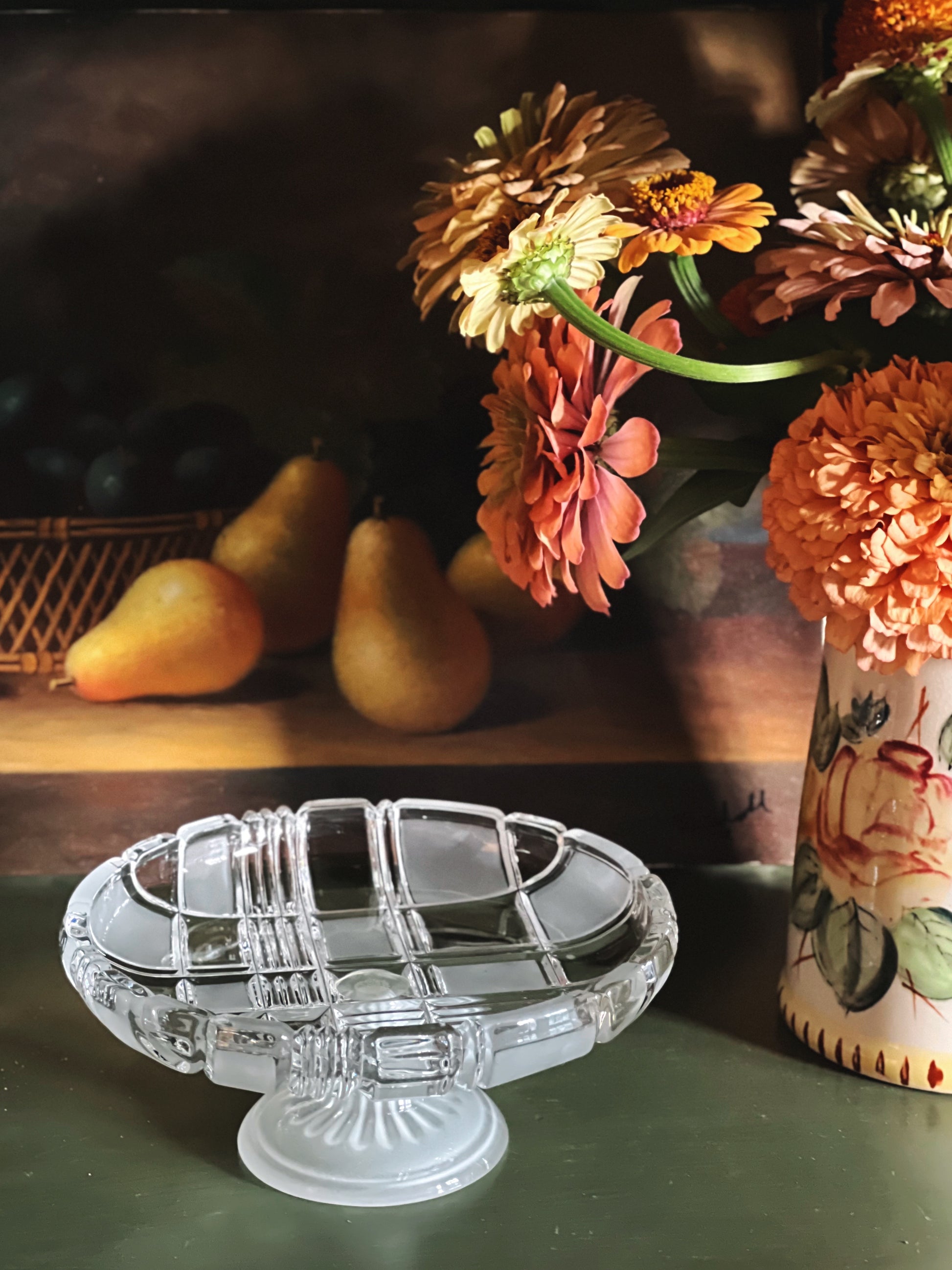 A clear glass pedestal plate with a ribbed base and a matching glass bowl, styled on a dark green surface with a floral vase of orange flowers and pears in the background.