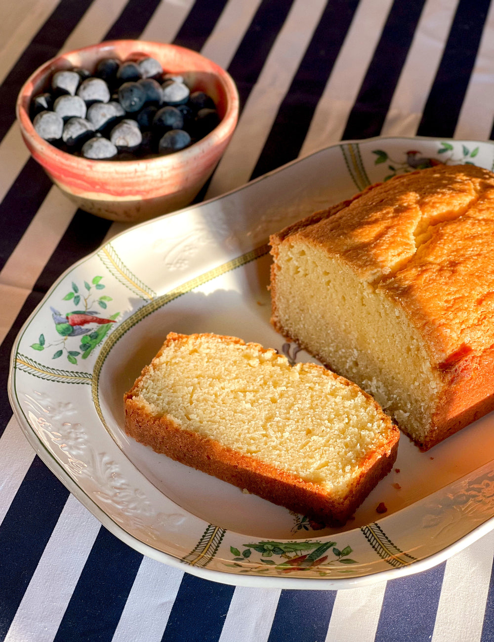 Classic pound cake on a vintage floral platter with fresh blueberries on a striped tablecloth.