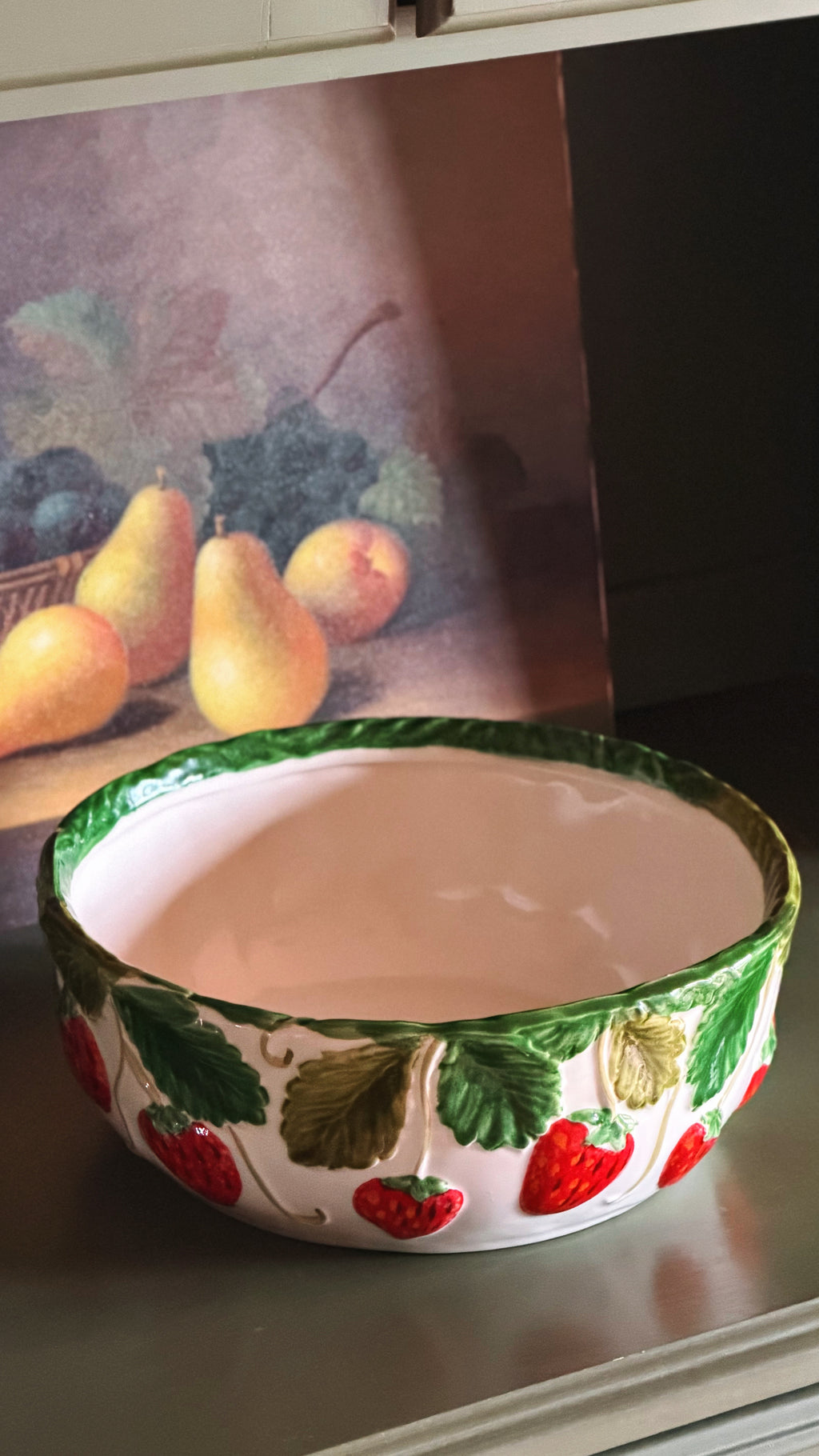 A ceramic bowl with a white interior and a decorated exterior featuring green leaves and red strawberries, set on a green surface with a fruit painting in the background.