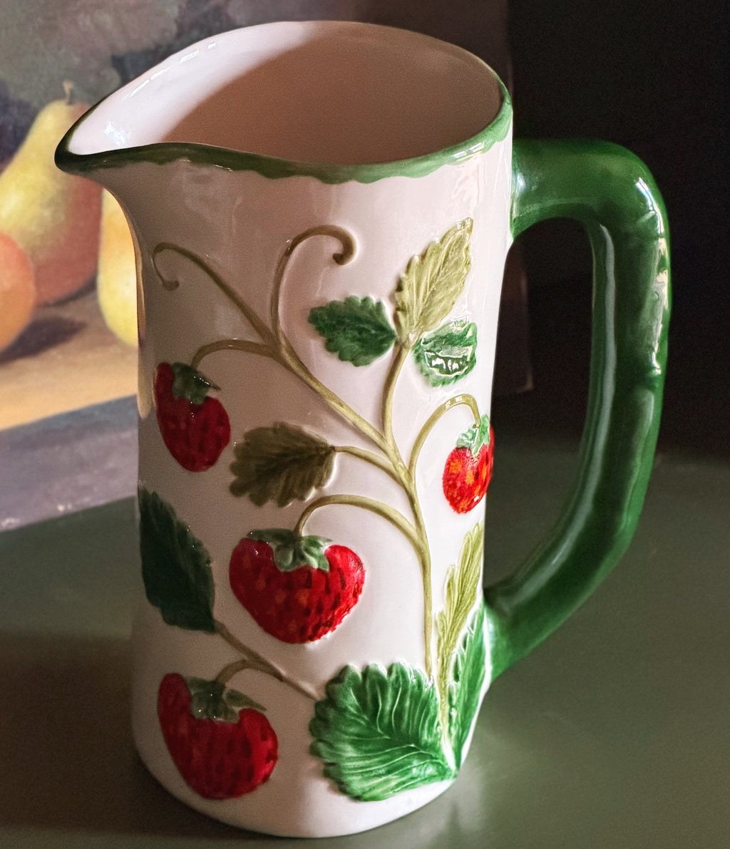 A white ceramic pitcher with a green handle and rim, decorated with red strawberries and green leaves on a floral vine pattern, set against a dark background.