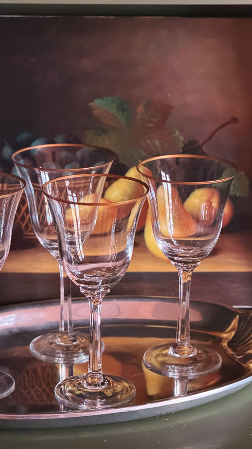 A set of Vintage Lenox Hayworth Crystal Wine Glasses with gold rims, arranged on a reflective silver tray against a dark, painterly background depicting fruit and leaves.