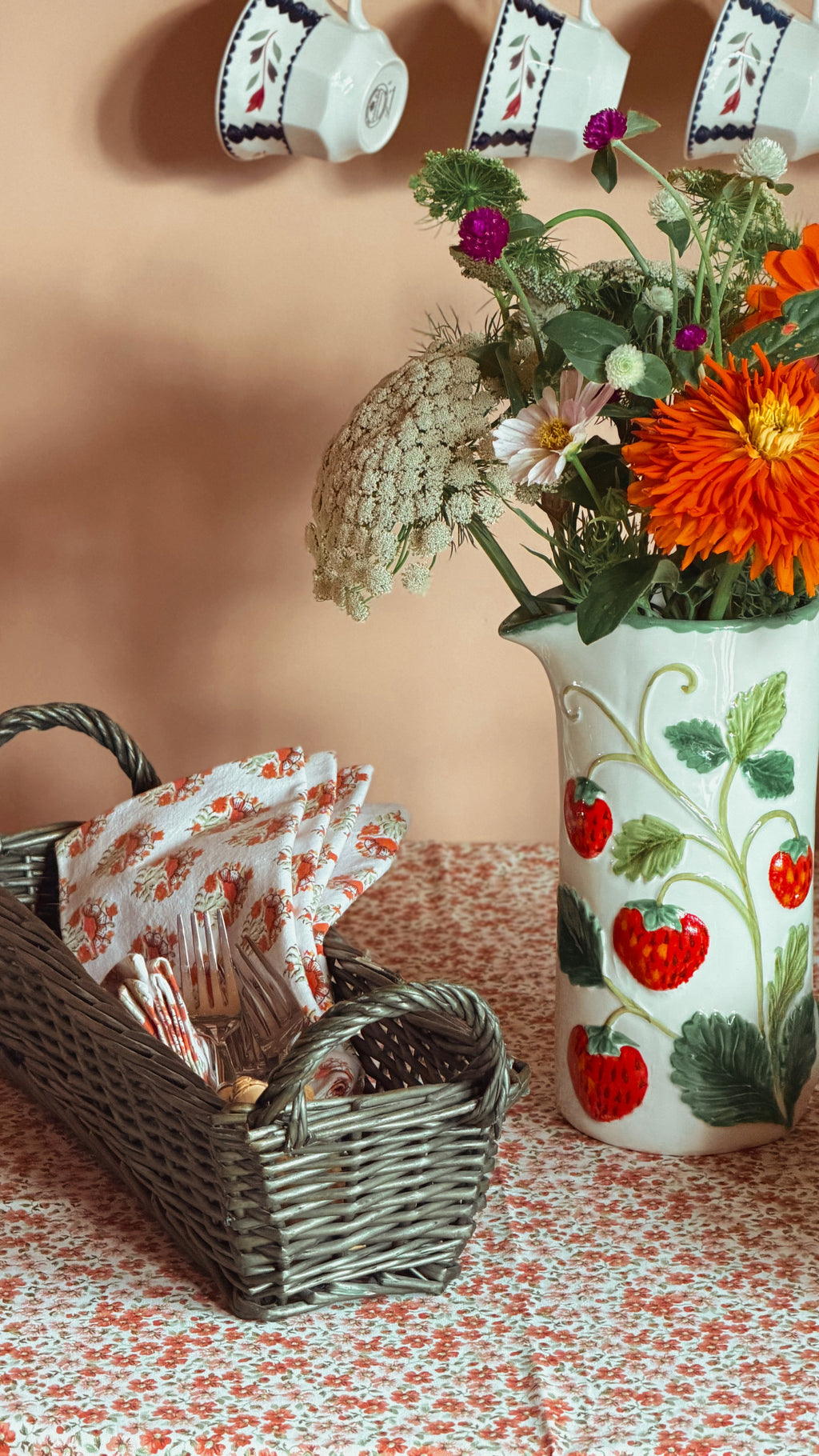A white ceramic pitcher decorated with red strawberries and green leaves, filled with a colorful flower bouquet, set on a floral tablecloth next to a vintage green wicker basket holding patterned napkins.