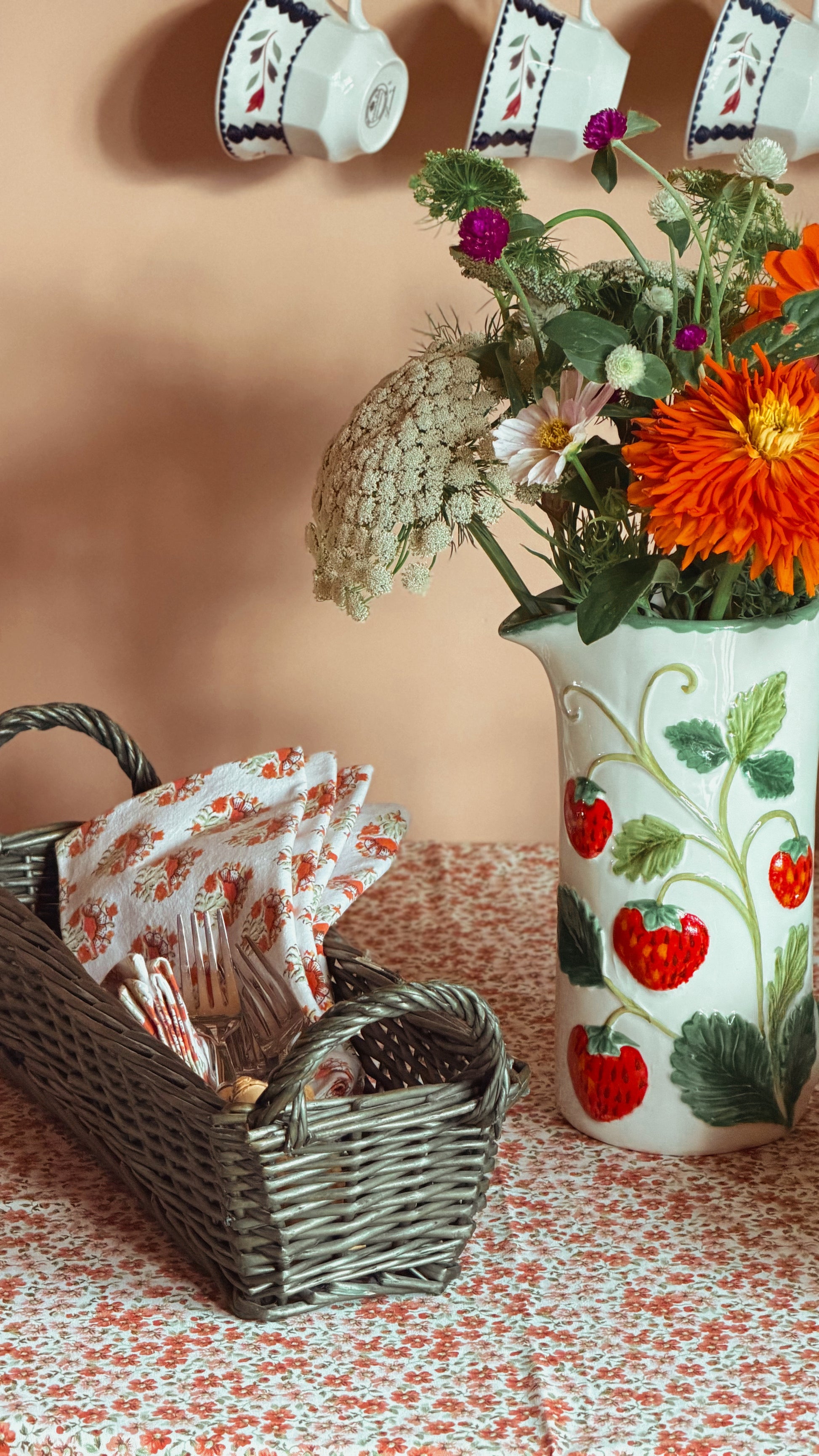 A white ceramic pitcher decorated with red strawberries and green leaves, filled with a colorful flower bouquet, set on a floral tablecloth next to a vintage green wicker basket holding patterned napkins.