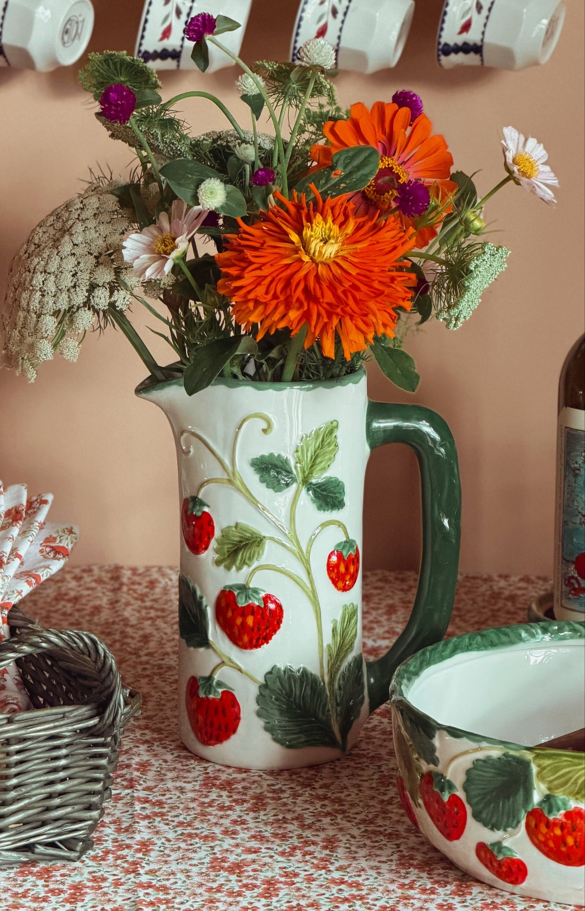A white ceramic pitcher and matching bowl decorated with red strawberries and green leaves, filled with a bouquet of orange, white, and purple flowers, set on a patterned tablecloth with hanging teacups in the background.