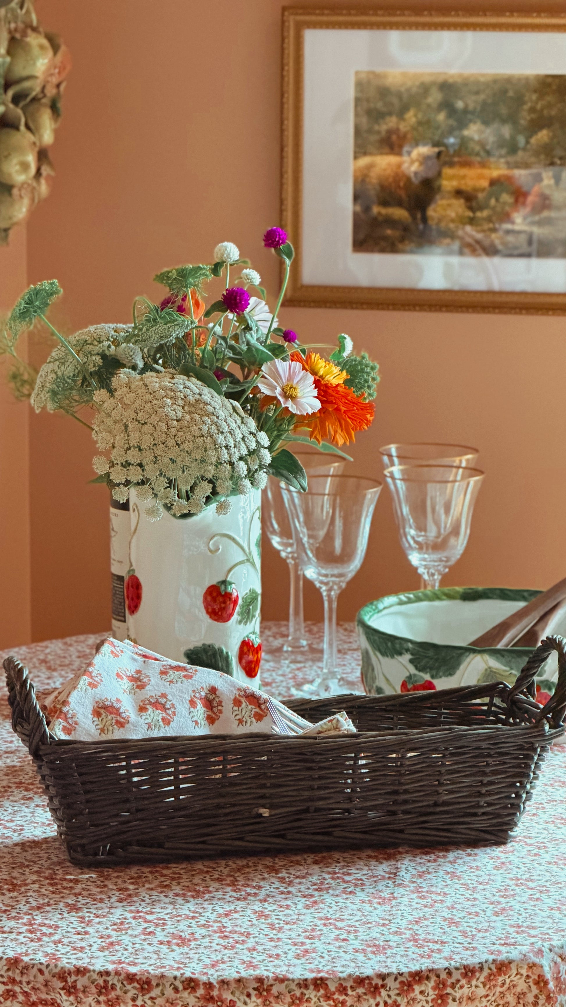 A styled tabletop scene with a white ceramic vase decorated with red strawberries holding a bouquet of mixed wildflowers, a Vintage Green Wicker Basket with a floral napkin, clear wine glasses, and a green-and-white bowl on a patterned tablecloth against a warm peach wall.