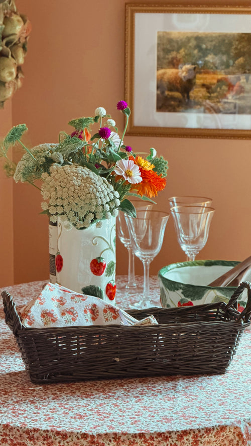 A styled tabletop scene with a white ceramic vase decorated with red strawberries holding a bouquet of mixed wildflowers, a Vintage Green Wicker Basket with a floral napkin, clear wine glasses, and a green-and-white bowl on a patterned tablecloth against a warm peach wall.