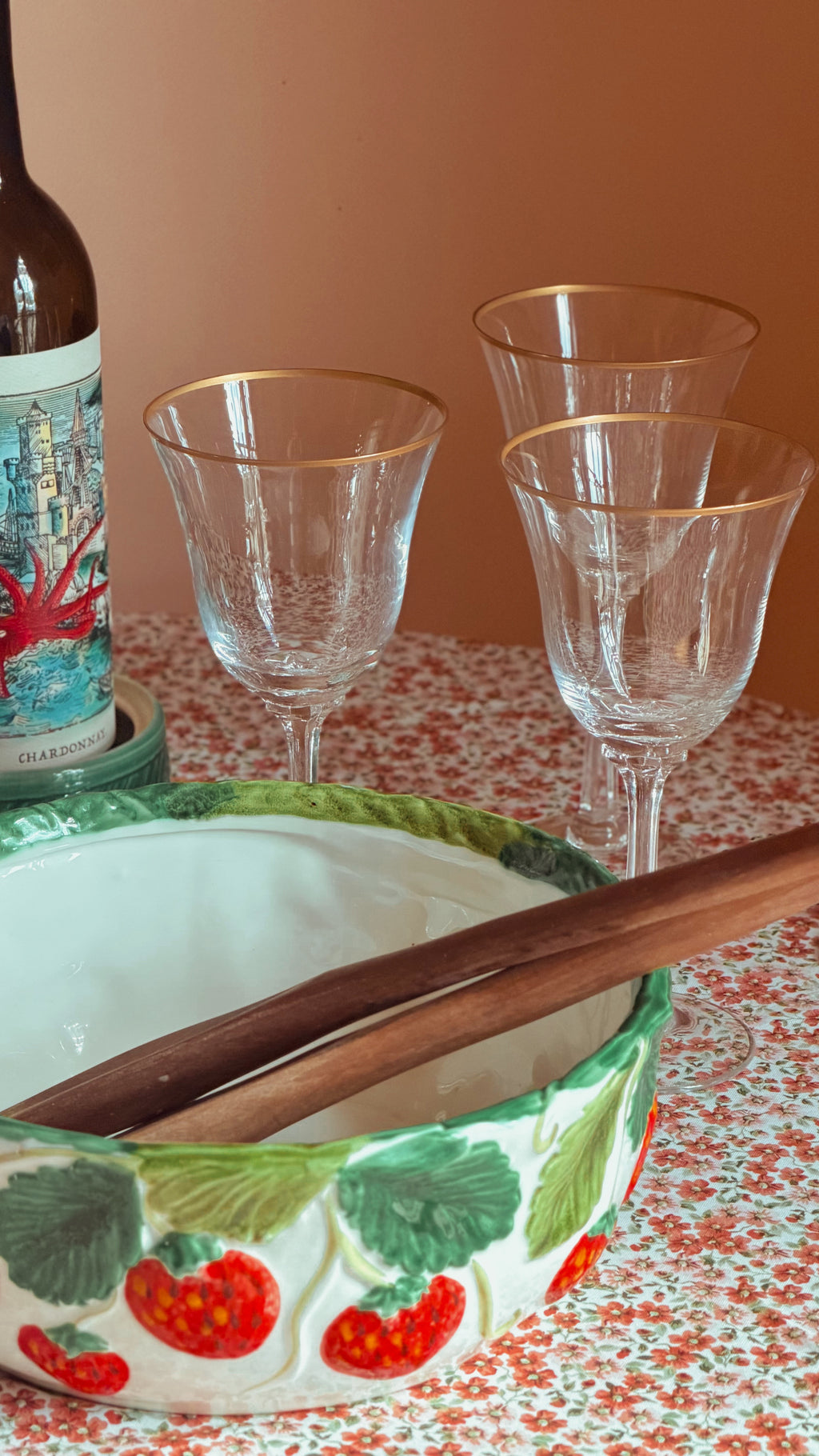 A close-up of a decorative ceramic bowl with a strawberry pattern, wooden spoons inside, three clear stemmed glasses with gold rims, and part of a wine bottle on a patterned tablecloth against a warm peach background.