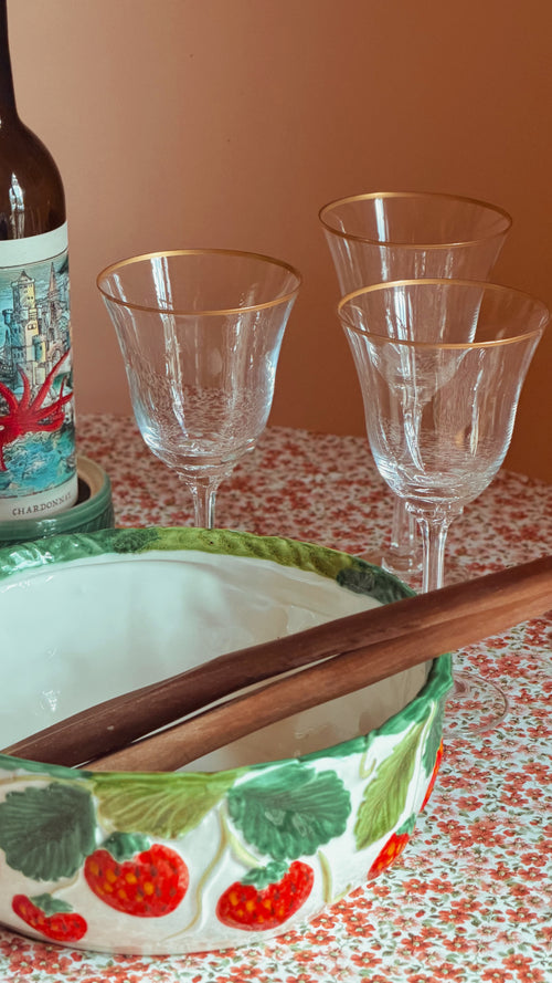 A close-up of a decorative ceramic bowl with a strawberry pattern, wooden spoons inside, three clear stemmed glasses with gold rims, and part of a wine bottle on a patterned tablecloth against a warm peach background.