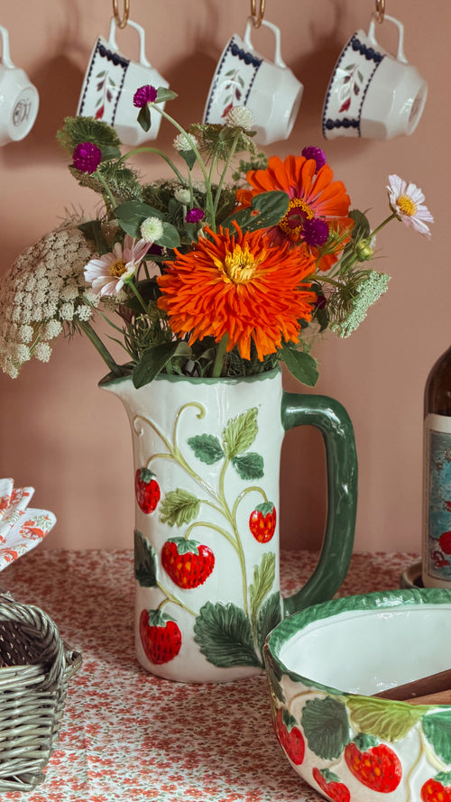 A ceramic pitcher vase decorated with red strawberries and green leaves, filled with a colorful wildflower bouquet, set on a patterned tablecloth with other matching strawberry-themed tableware in a warm, rustic setting.