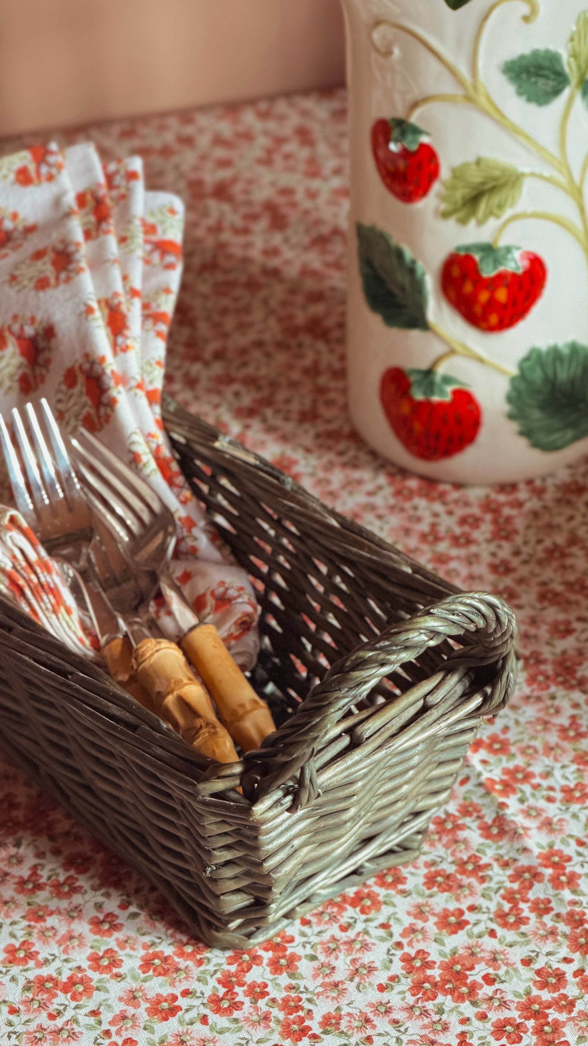 A small woven basket holding silver forks and wooden-handled utensils, placed on a red-and-white floral tablecloth beside a white ceramic pitcher decorated with red strawberries and green leaves.