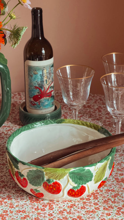 A decorative ceramic bowl with a strawberry pattern, containing wooden serving utensils, set on a floral tablecloth next to a wine bottle and clear glasses.