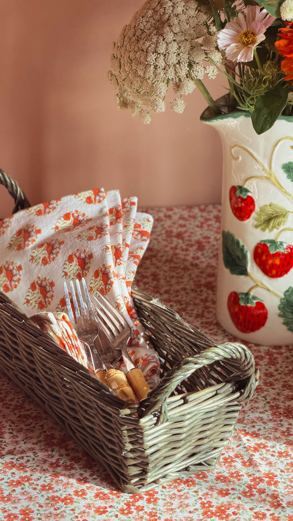 A vintage green wicker basket filled with a floral napkin, forks, and  placed on a matching floral tablecloth beside a strawberry-patterned ceramic vase with flowers.