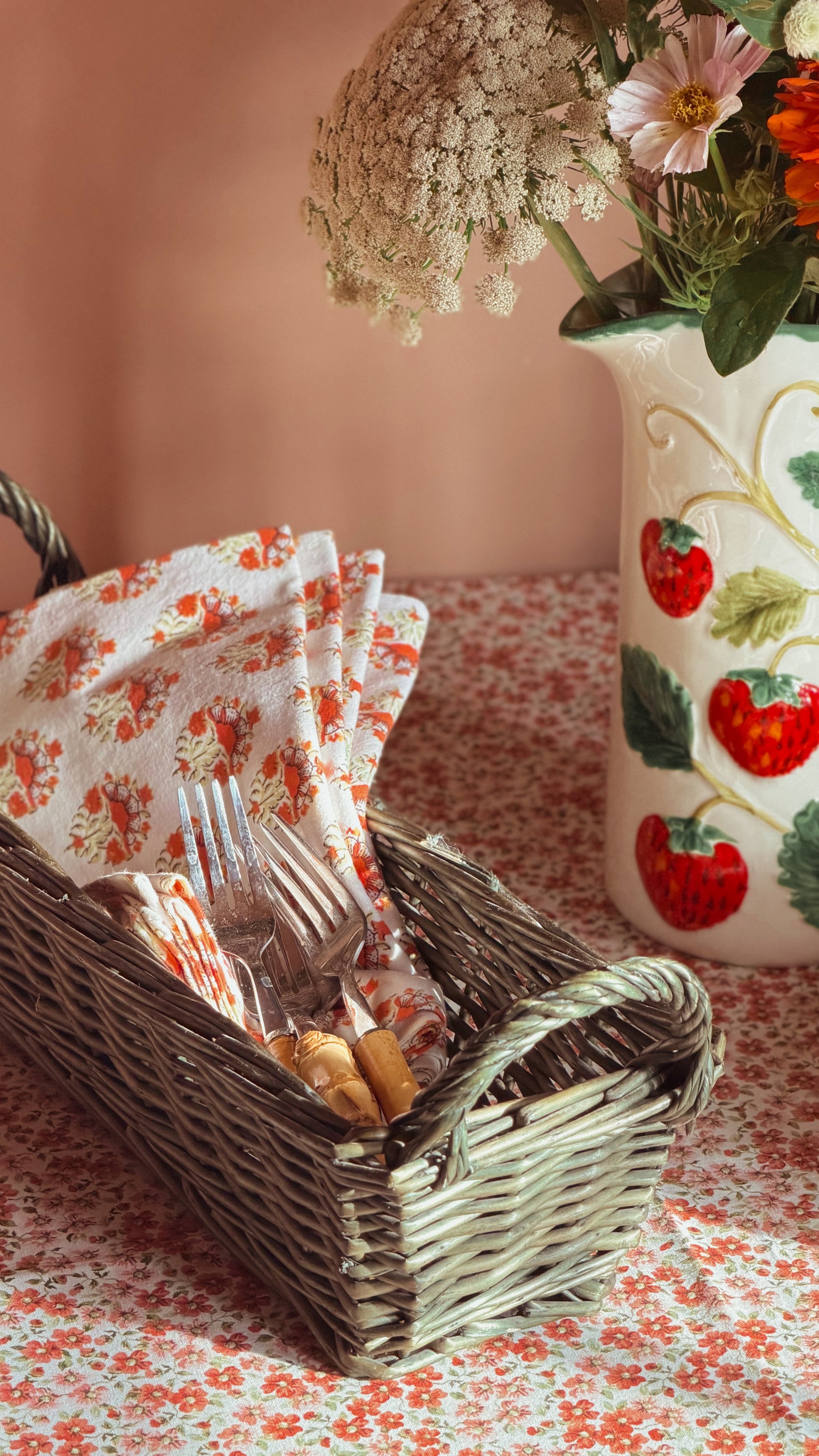 A vintage green wicker basket filled with a floral napkin, forks, and  placed on a matching floral tablecloth beside a strawberry-patterned ceramic vase with flowers.
