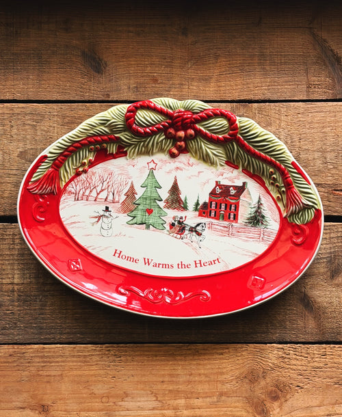A festive oval ceramic platter with a bright red rim and a raised holiday wreath design at the top. The center features a white winter village scene with a Christmas tree, snowman, house, sleigh, and the text 'Home Warms the Heart.