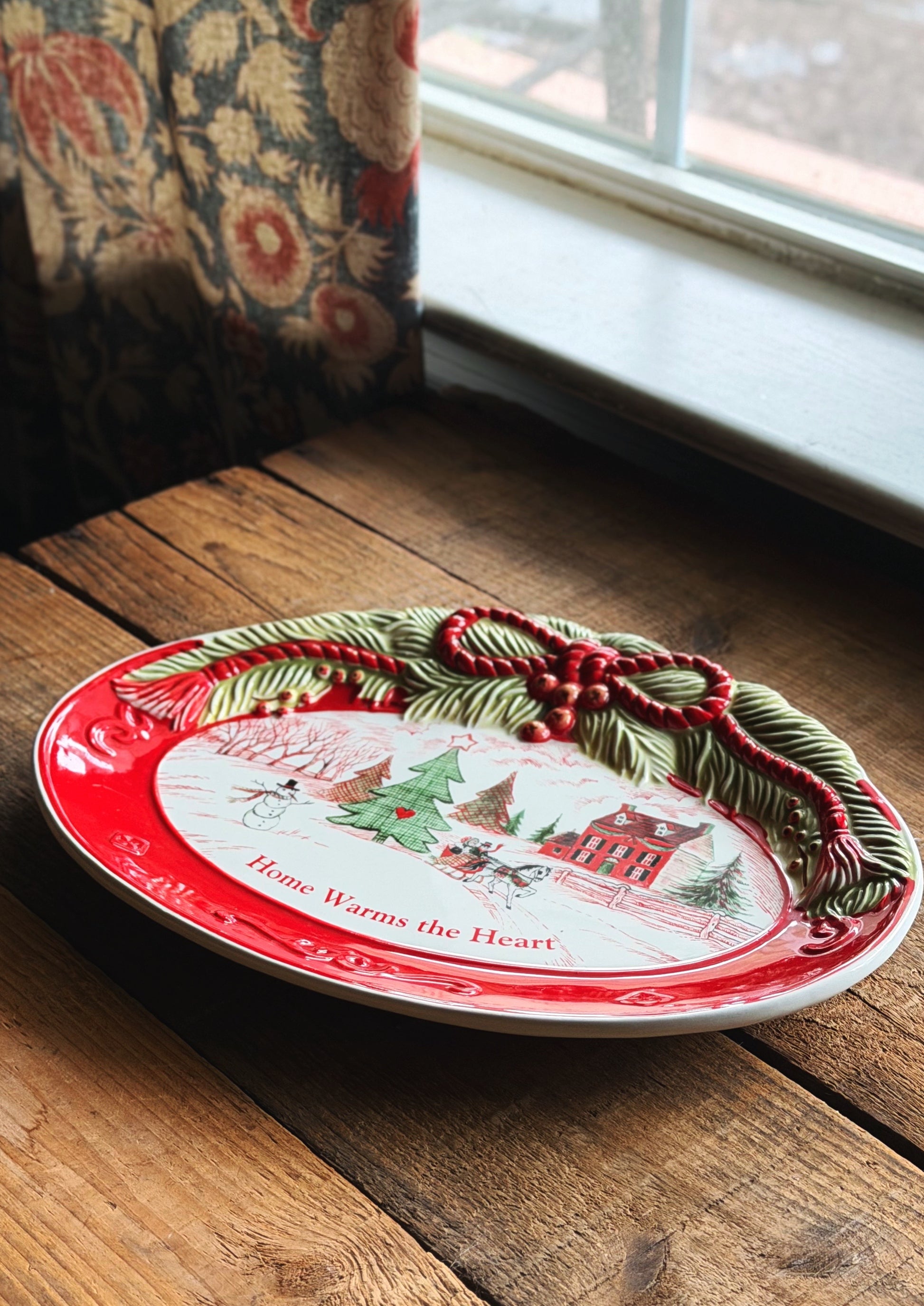 Vintage Fitz & Floyd holiday sentiment tray  with a red rim, holiday winter scene illustration, and 3D green pine garland with red berries, resting on a rustic wooden table near a window.