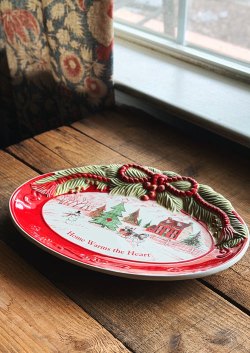 Vintage Fitz & Floyd holiday sentiment tray  with a red rim, holiday winter scene illustration, and 3D green pine garland with red berries, resting on a rustic wooden table near a window.