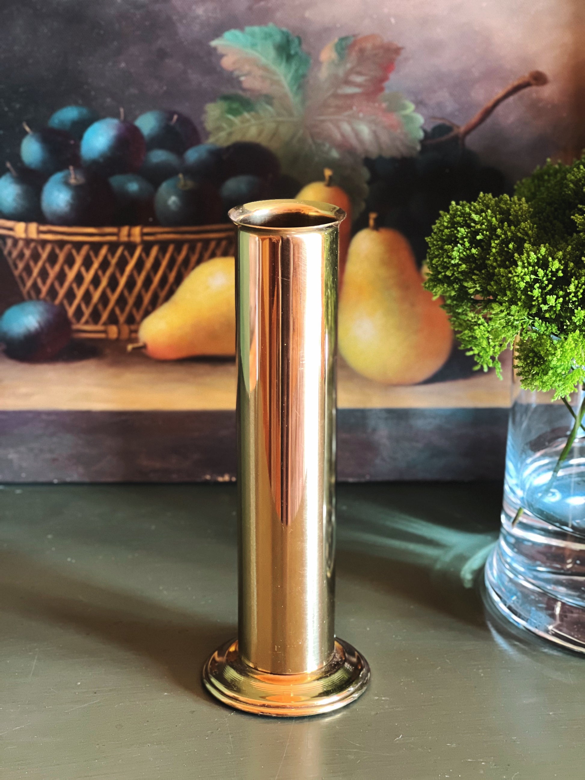 A tall, cylindrical brass candlestick holder or vase with a polished metallic finish, standing on a dark surface with a rustic still life background depicting pears and grapes.