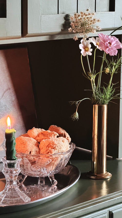 A styled interior scene with a glass bowl of sponges on a silver tray beside a lit green candle, next to a Vintage Brass Cylinder Bud Vase  holding pink and white flowers against a dark wall and cabinet background.