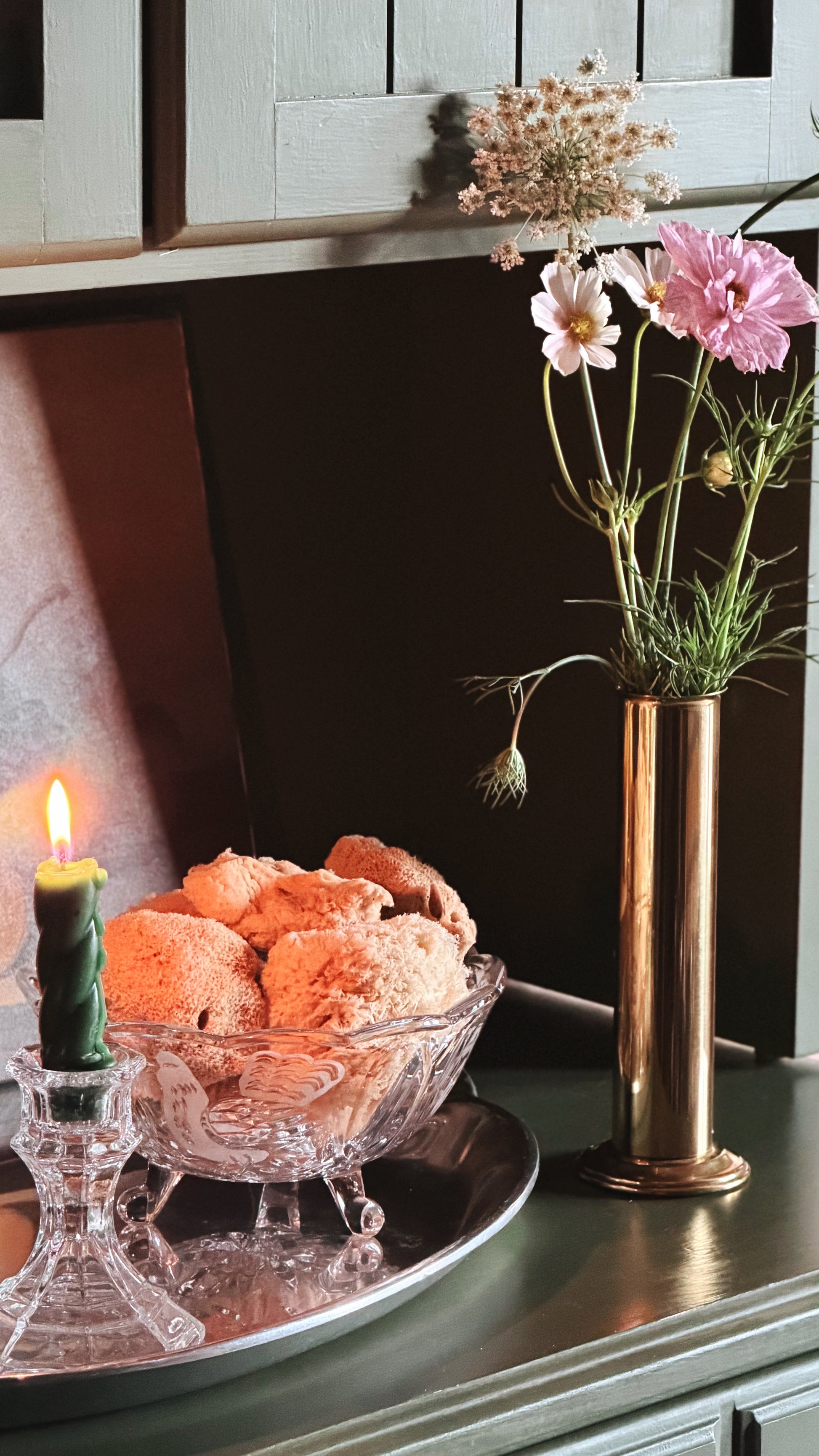 A styled interior scene with a glass bowl of sea sponges on a silver tray beside a lit green candle, next to a tall metallic vase holding pink and white flowers against a dark wall and cabinet background.