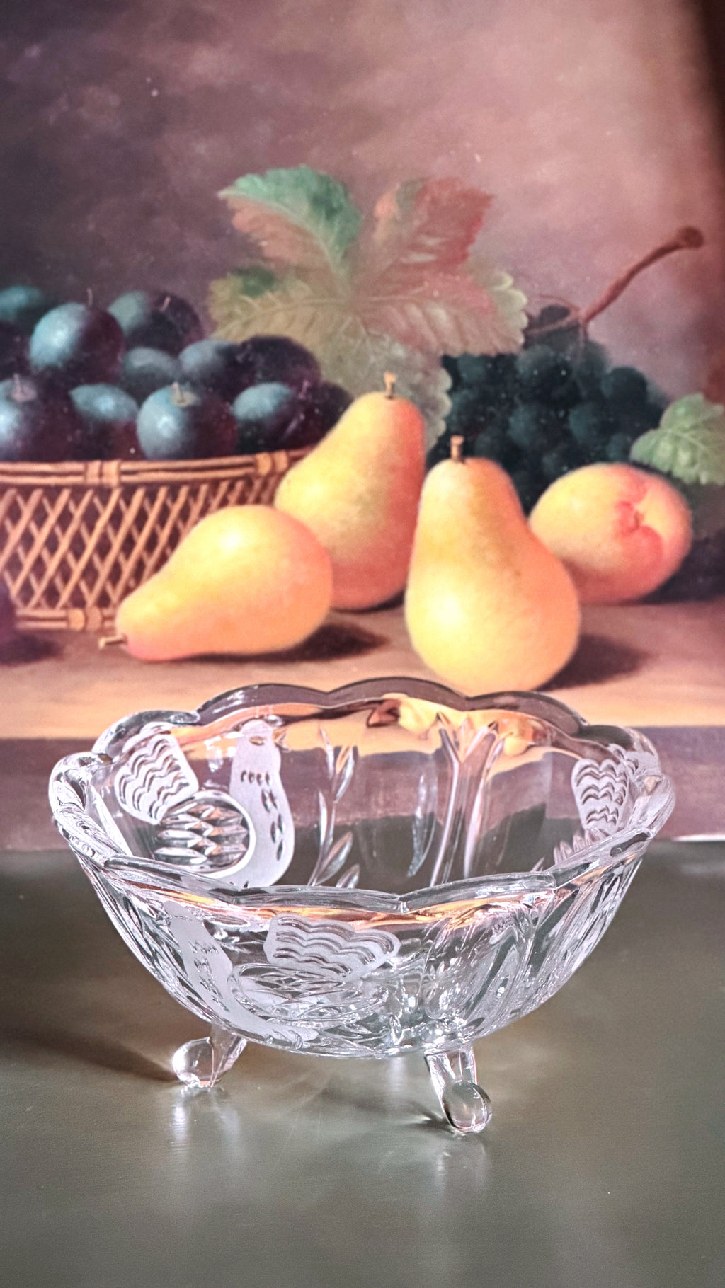 Vintage Crystal Footed Bowl with Etched Bird Design, placed in front of a painted still life background showing pears, grapes, and a basket of dark fruit.