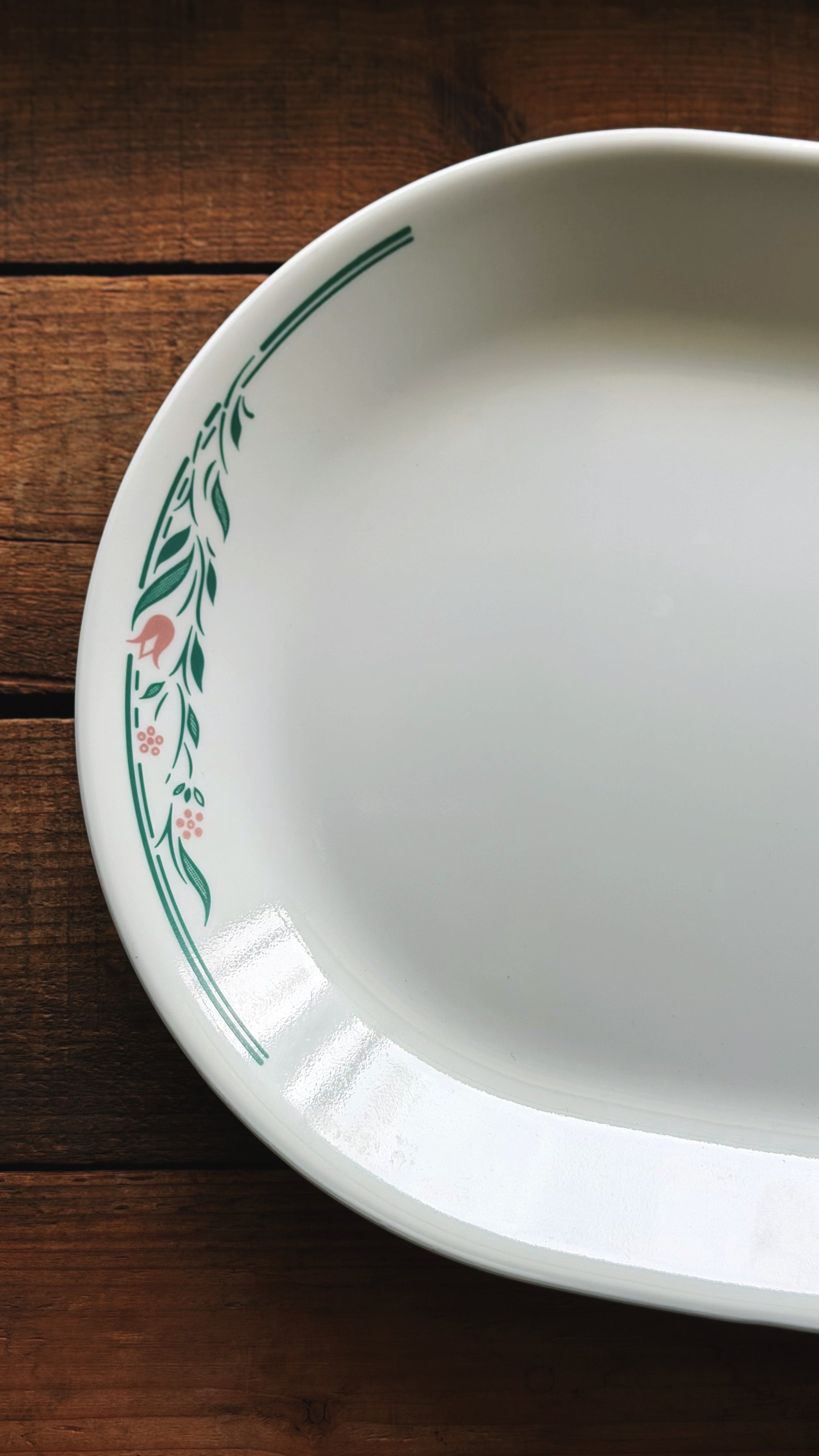 A Vintage Corelle Rosemarie Oval Serving Platter with a green leafy border and small pink floral accents, shown on a dark wooden surface.