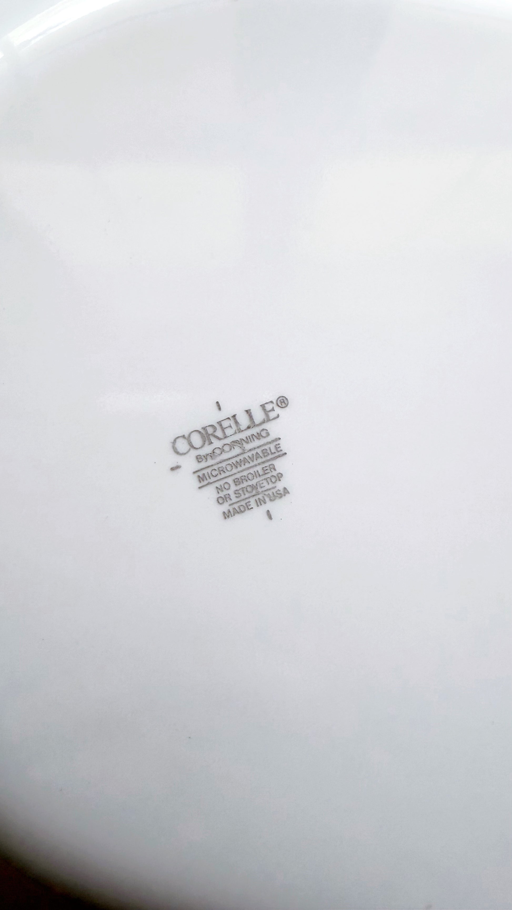 Close-up of the bottom of a white Corelle ceramic plate or dish, showing the molded Corelle brand mark and small printed text.