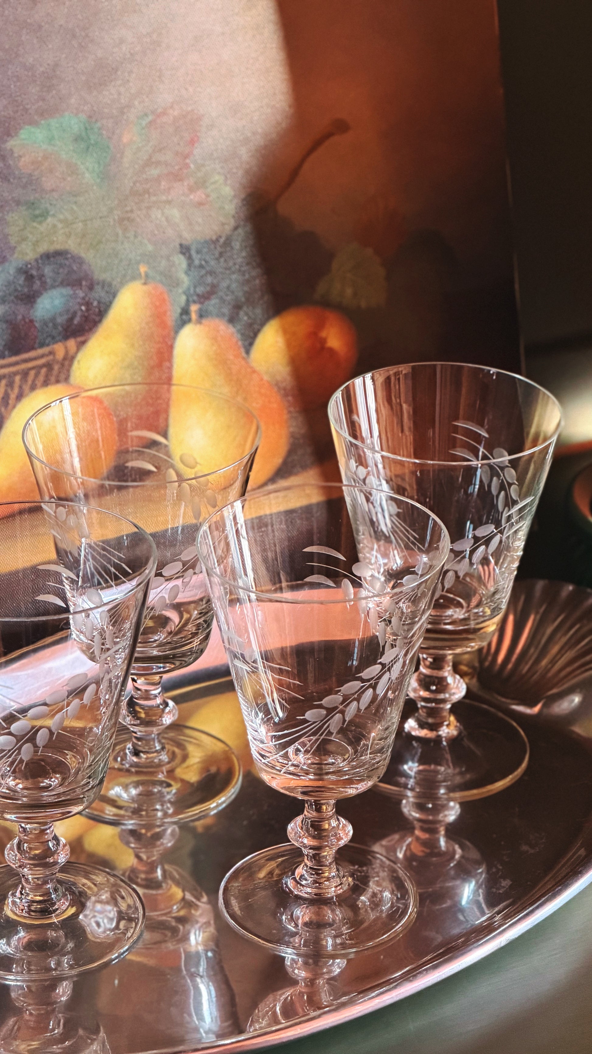A set of wine glasses with etched floral patterns, arranged on a reflective silver tray in warm, dim lighting with a pear painting in the background.