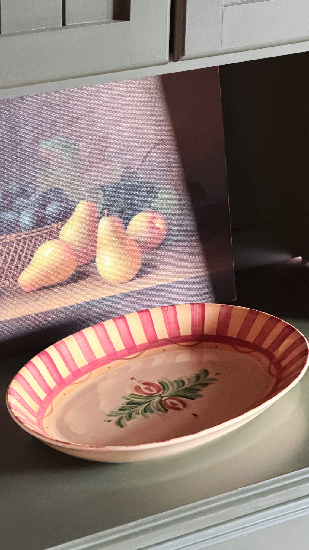 A Vintage Gail Pittman Siena Serving Platter displayed on a shelf in front of a still life fruit painting.